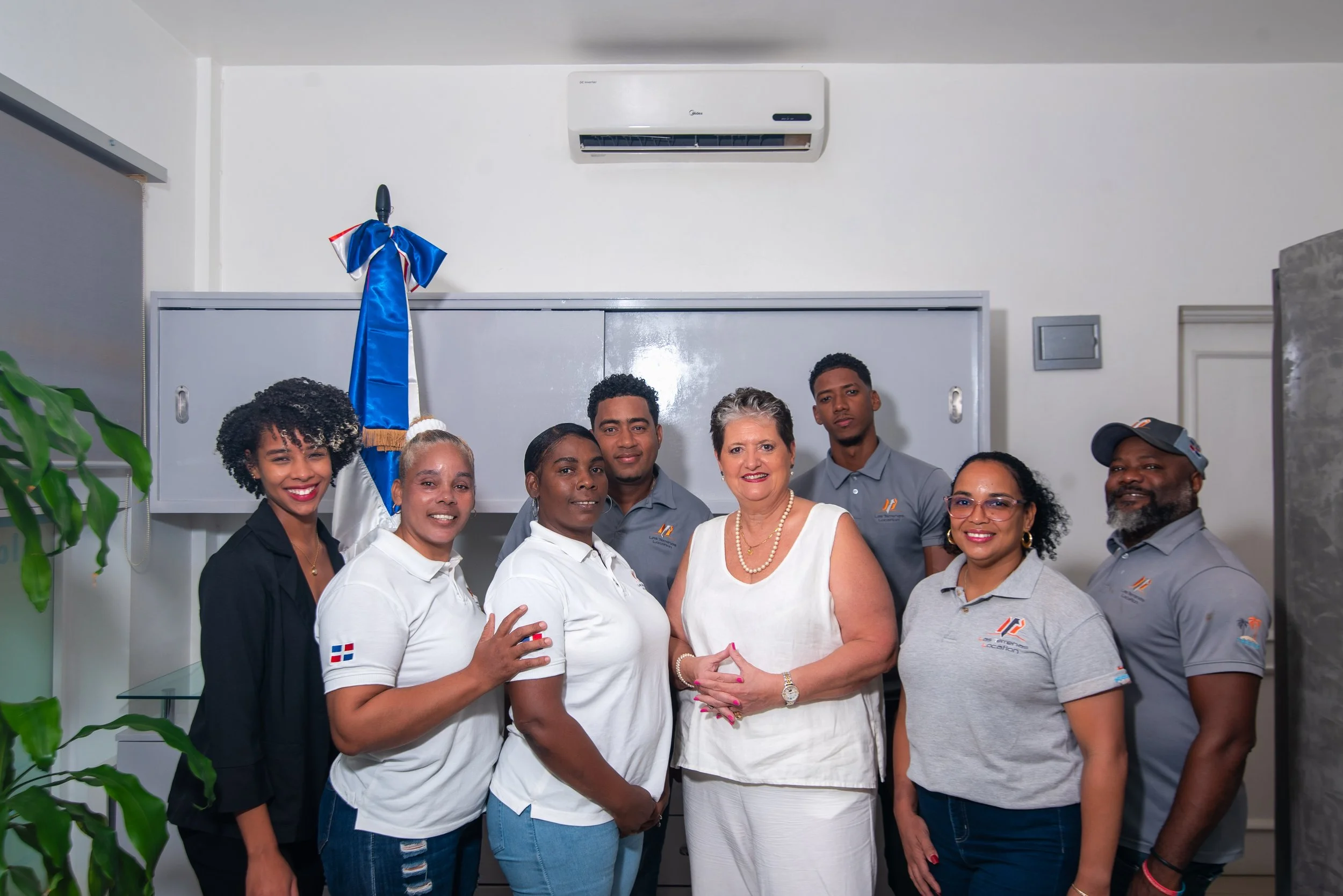 Grupo de ocho personas posando en una oficina con bandera y aire acondicionado.