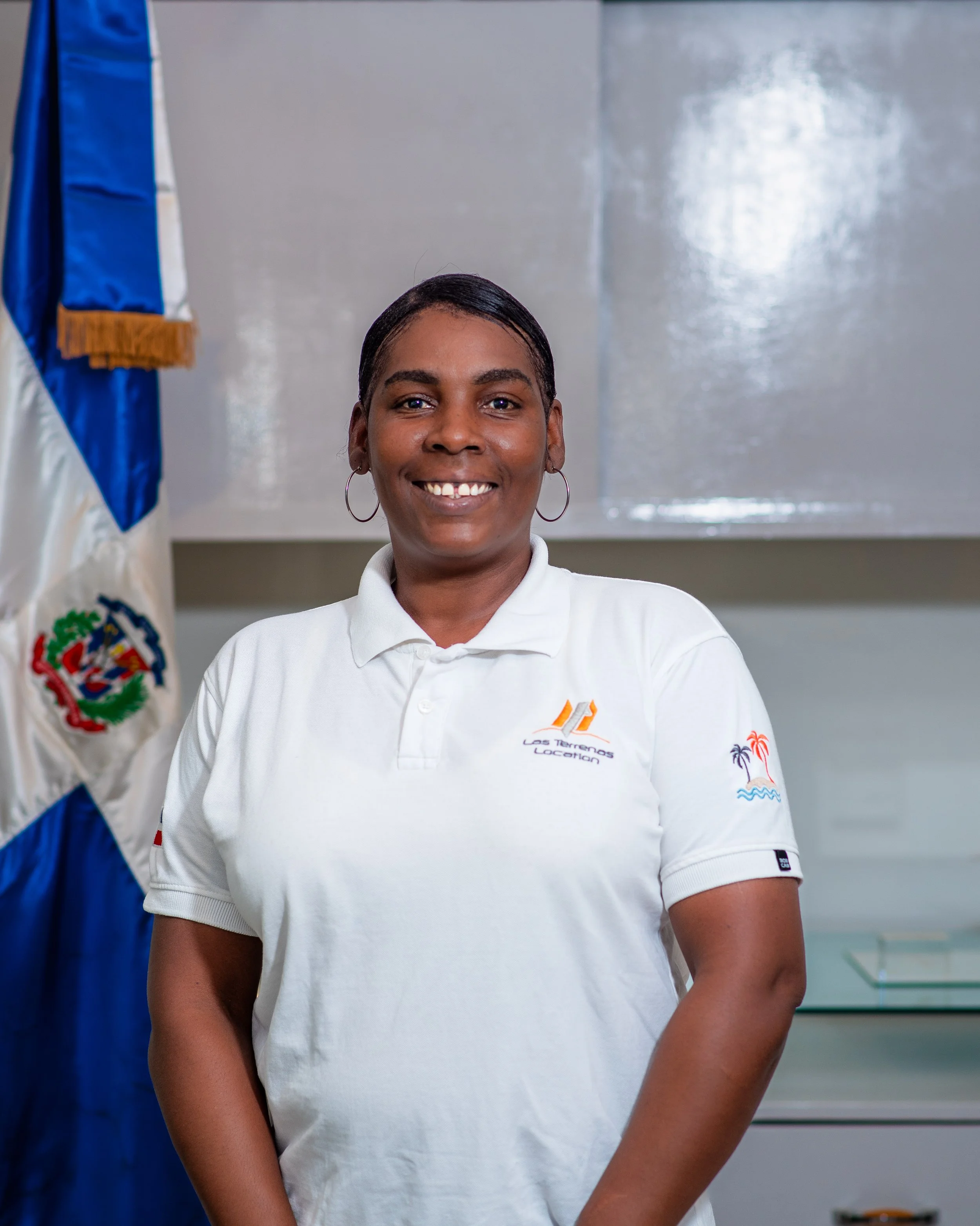 Mujer sonriente con camiseta blanca y aretes, frente a bandera dominicana.