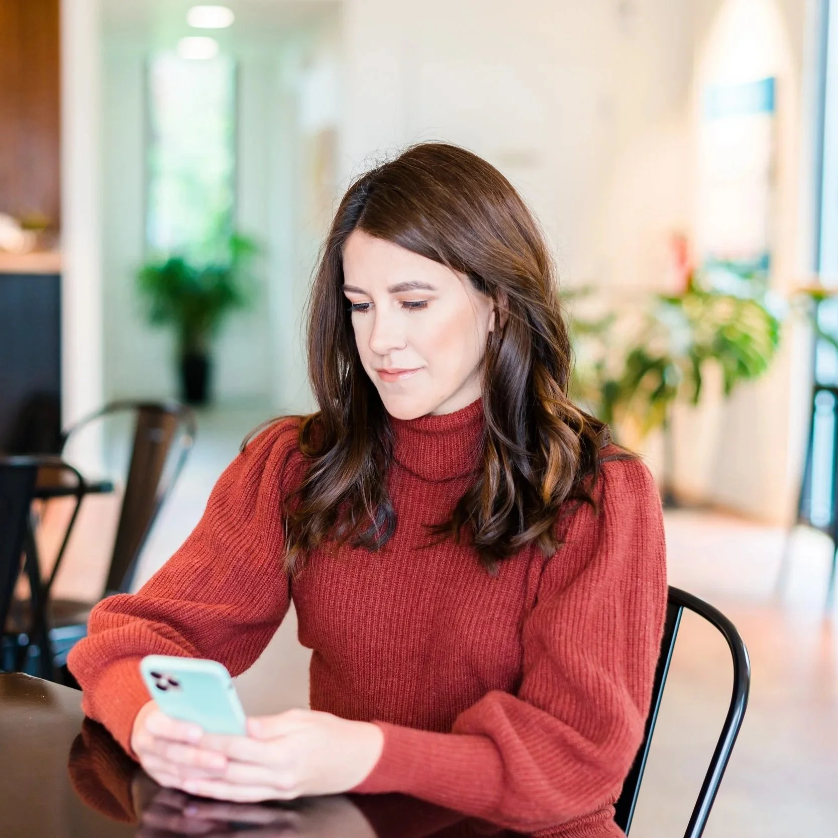 A young woman with long brown hair wearing a red sweater sitting at a table in a cozy cafe, looking at her smartphone.