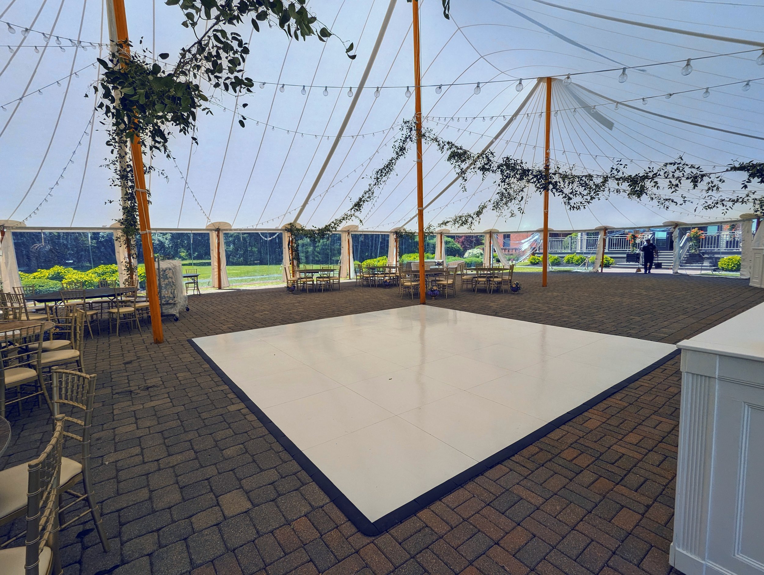 Elegant banquet hall set up for a wedding reception with round and rectangular tables, floral centerpieces, place settings, a DJ booth, string lights, large windows offering a scenic outdoor view, and a stacked wedding cake by the window.