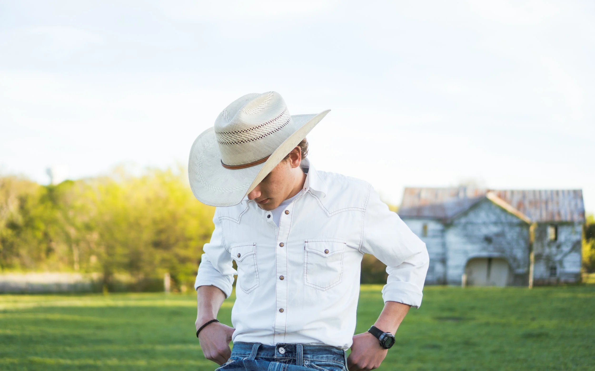 A young man wearing a white cowboy hat, white button-up shirt, and jeans standing outdoors with his hands on his hips, looking downward, with a blurred green landscape and an old barn in the background.