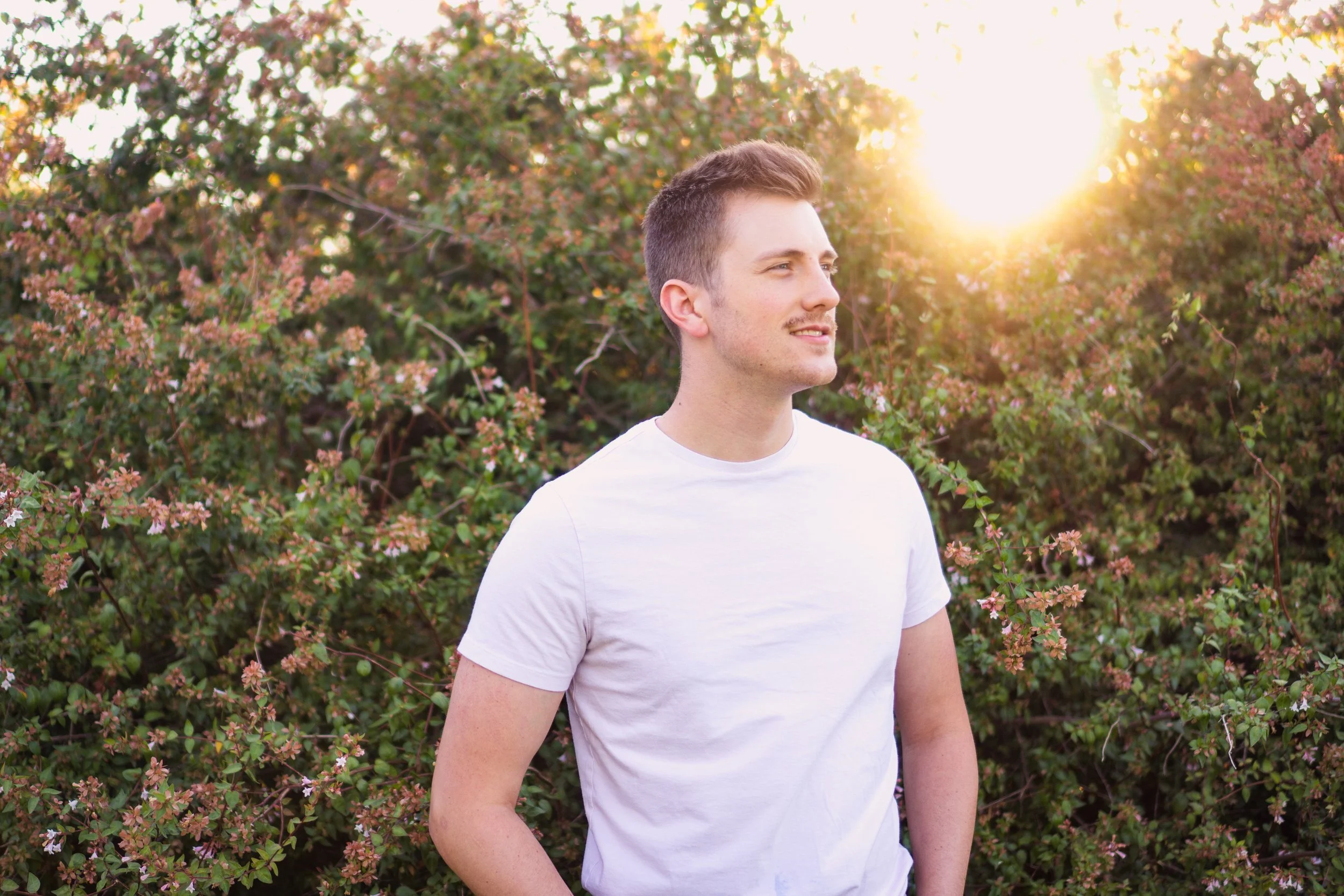 A young man with short brown hair and a light beard stands outdoors in front of a shrubbery with pink flowers, smiling slightly as the sun sets behind him.