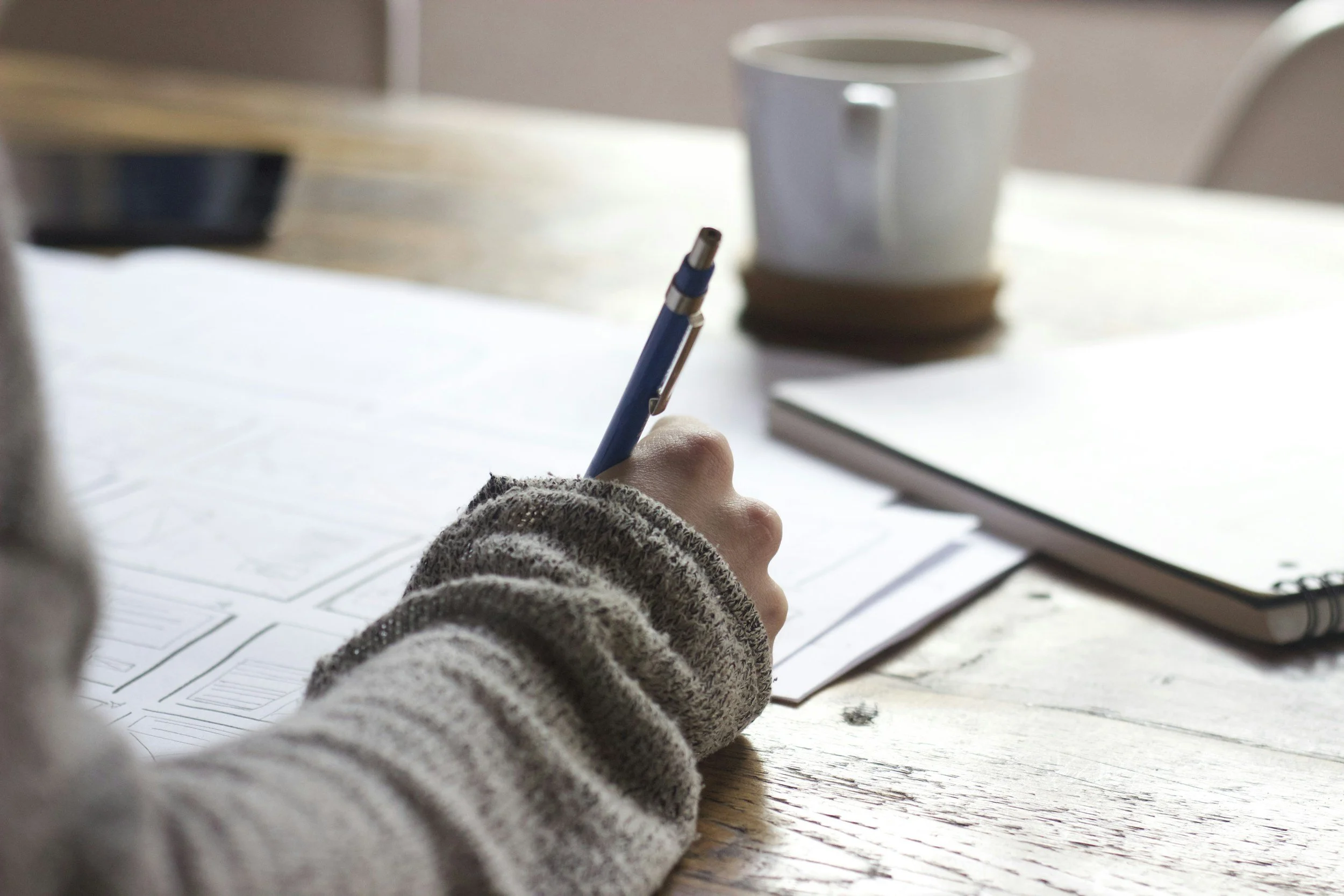 Person writing with a pen on paper at a wooden table with a cup and notebook nearby.