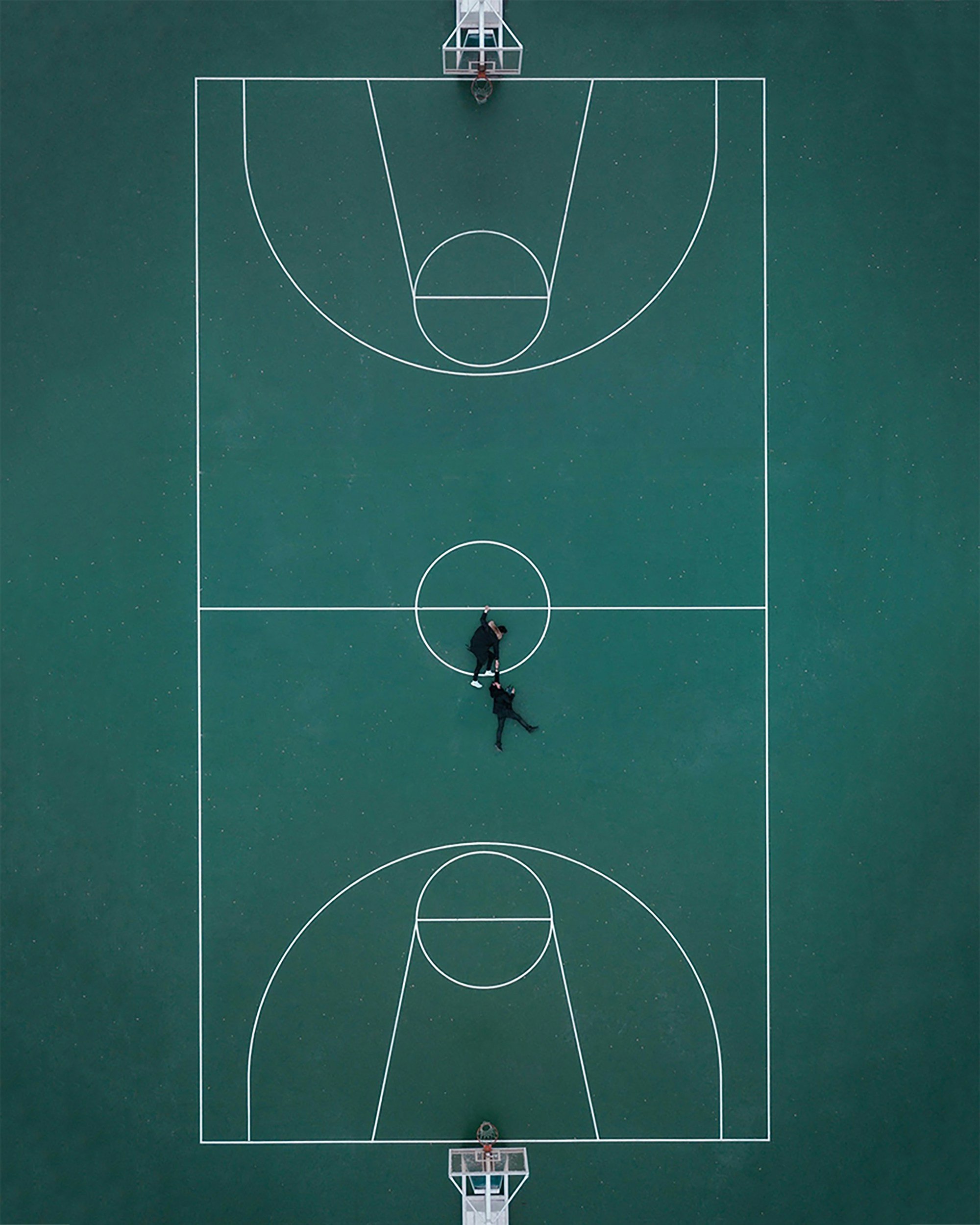 Aerial view of a basketball court with two people playing, basketball hoops at both ends, and court lines visible on green surface.