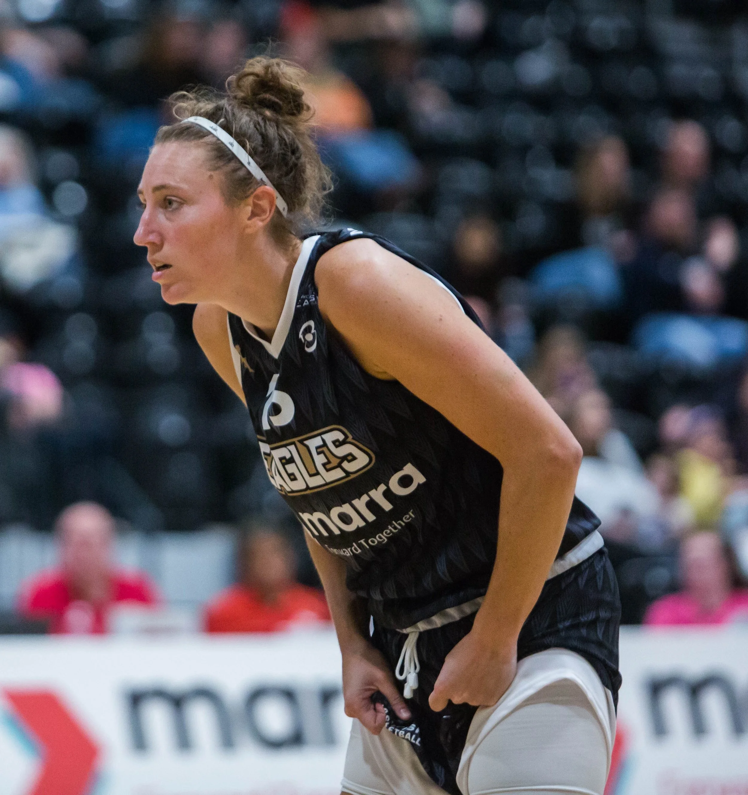 A female basketball player in a black and white uniform during a game, leaning forward with hands on her knees, focusing on the game.