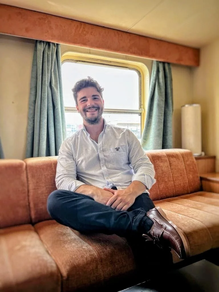 Homem sorridente sentado em sofá vintage com cortinas verdes e janela ao fundo, dentro de um ambiente aconchegante.