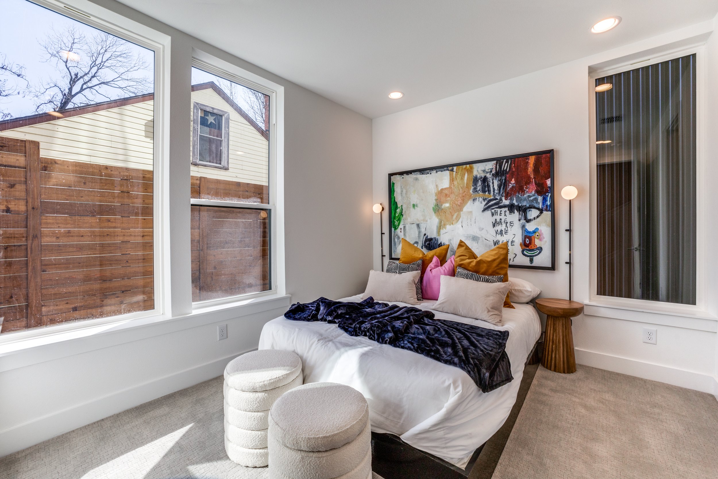 Modern bedroom with a large bed, colorful pillows, decorative artwork above the headboard, and two round stools. Large windows provide natural light, uncovering a view of a wooden fence and small building outside.