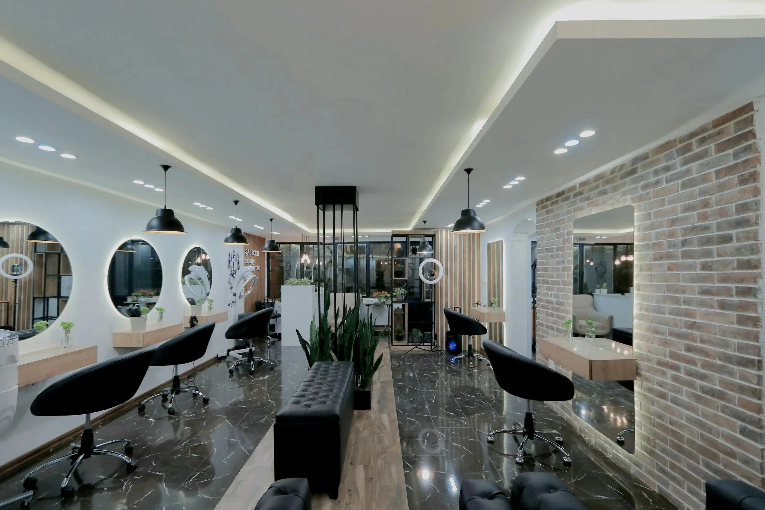 Interior of a modern hair salon with black chairs, round mirrors, wall decor, and a mix of white and brick walls, illuminated by ceiling lights and pendant lamps.