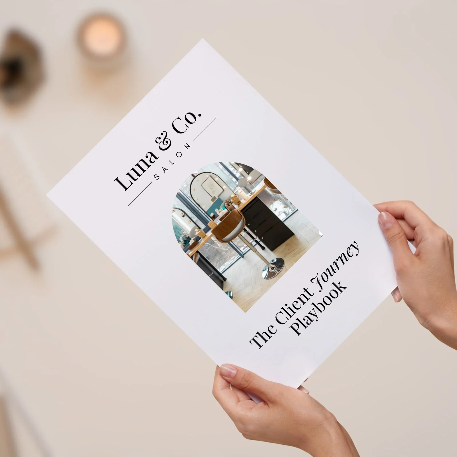 A person holds a printed booklet titled "Luna & Co. Salon" with a circular photo of a salon interior featuring chairs, mirrors, and a window. The booklet's subtitle reads "The Client Journey Playbook."