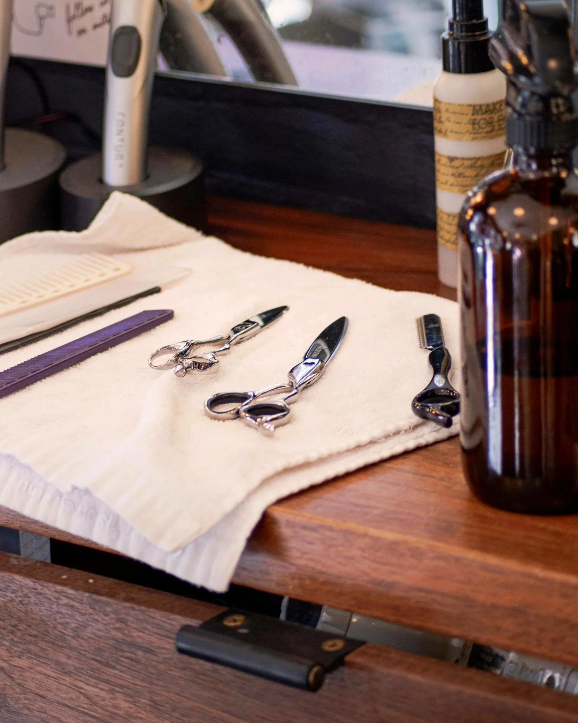 Hairdressing tools including scissors, thinning shears, a razor, and a comb placed on a white towel on a wooden countertop, with bottles and other hairdressing supplies in the background.