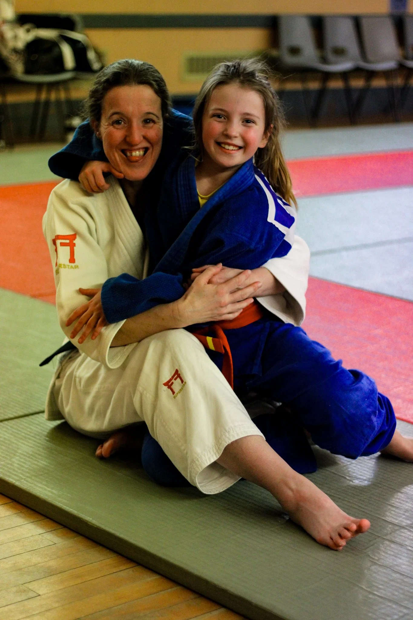 Two women in judo gi uniforms smiling and hugging on a mat in a dojo.