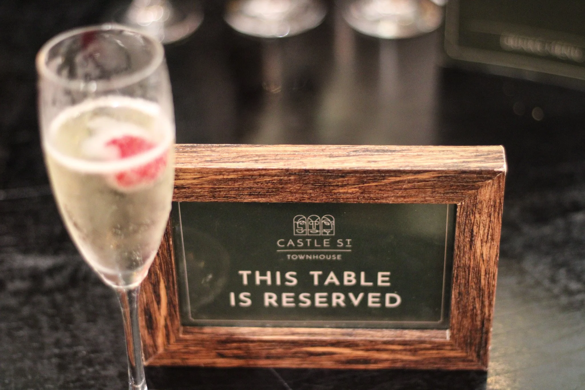 A glass of champagne with a raspberry and lemon wedge next to a wooden reserved sign on a dark table.