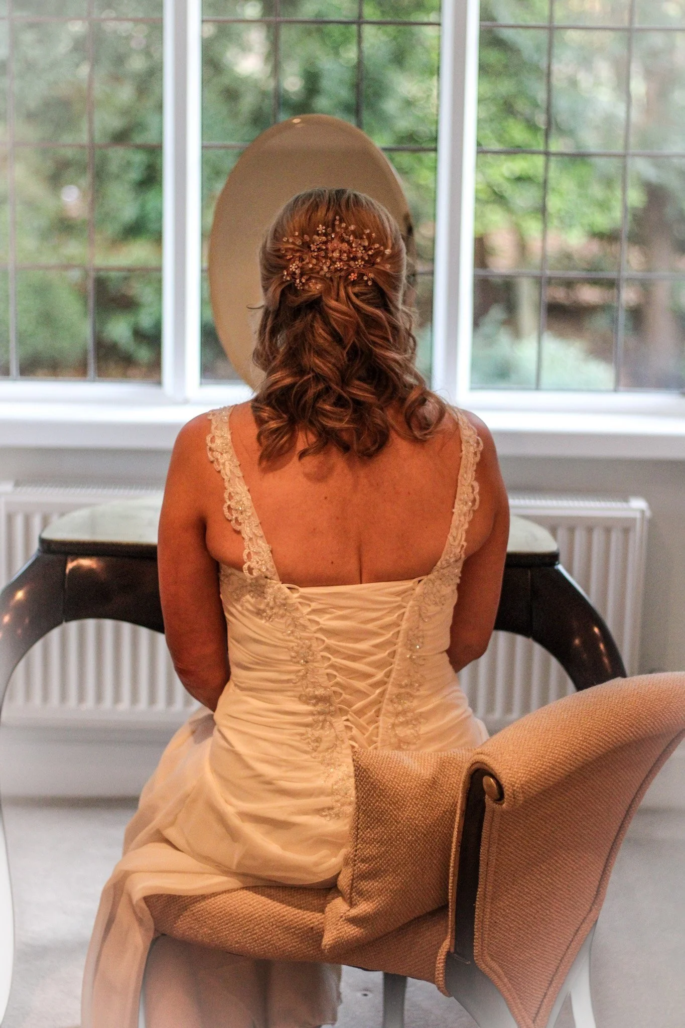 Woman seated in a dress facing a mirror in front of a window with greenery outside.