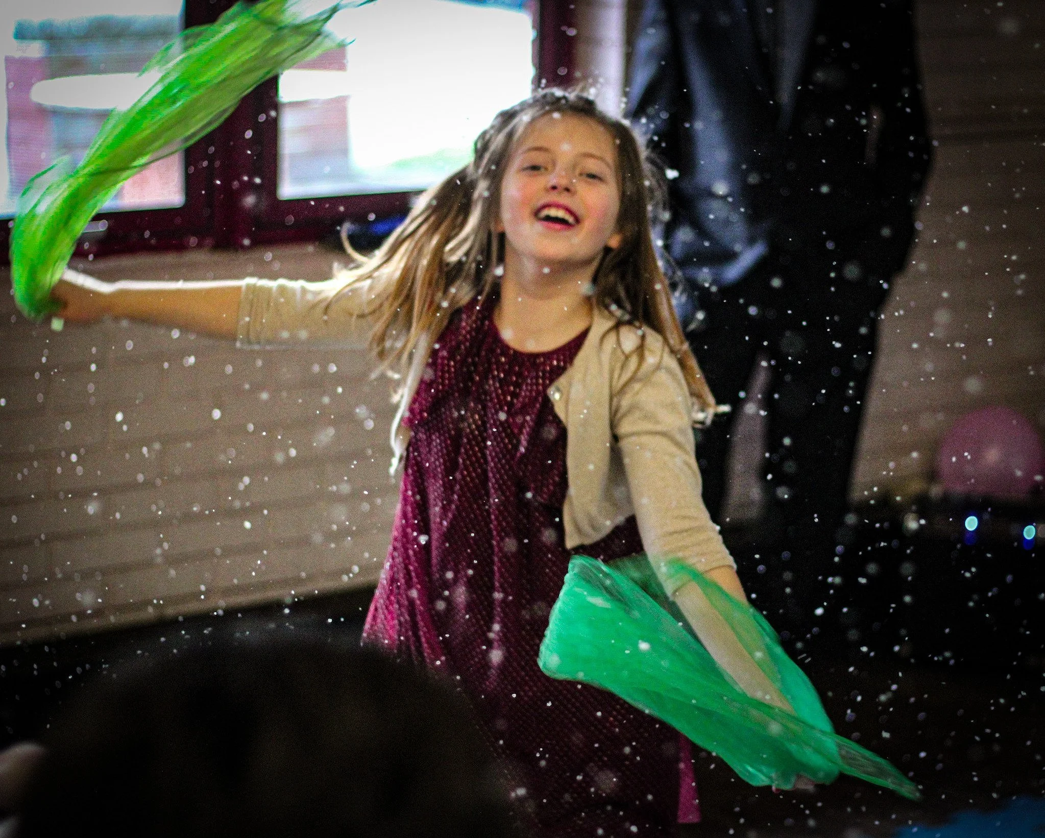 A young girl happily playing with green and yellow scarves, tossing them in the air with water droplets around her, indoors near a window.
