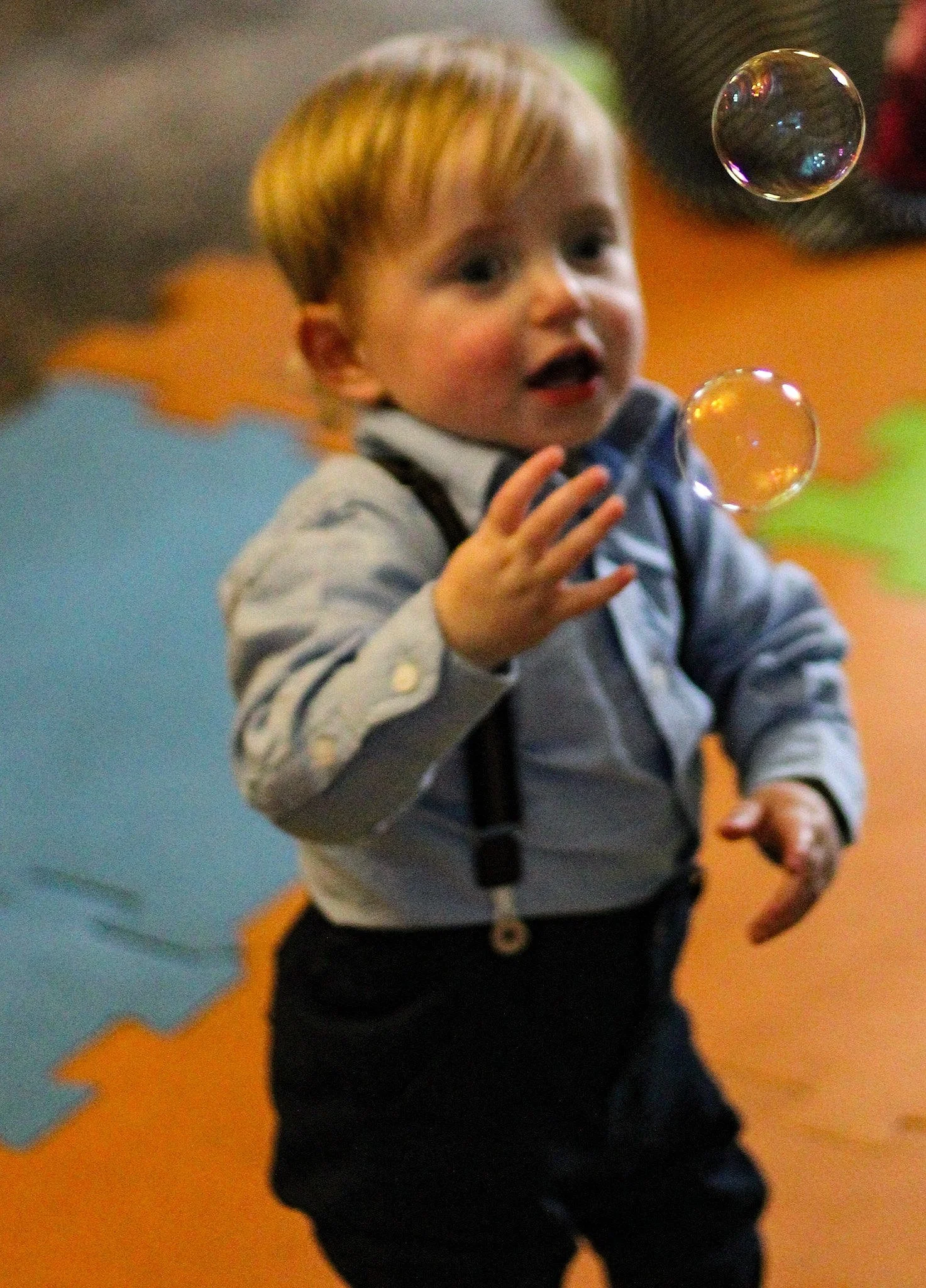 A young boy with blond hair wearing a gray shirt with suspenders, standing on colorful foam mats, reaching out to touch floating soap bubbles.