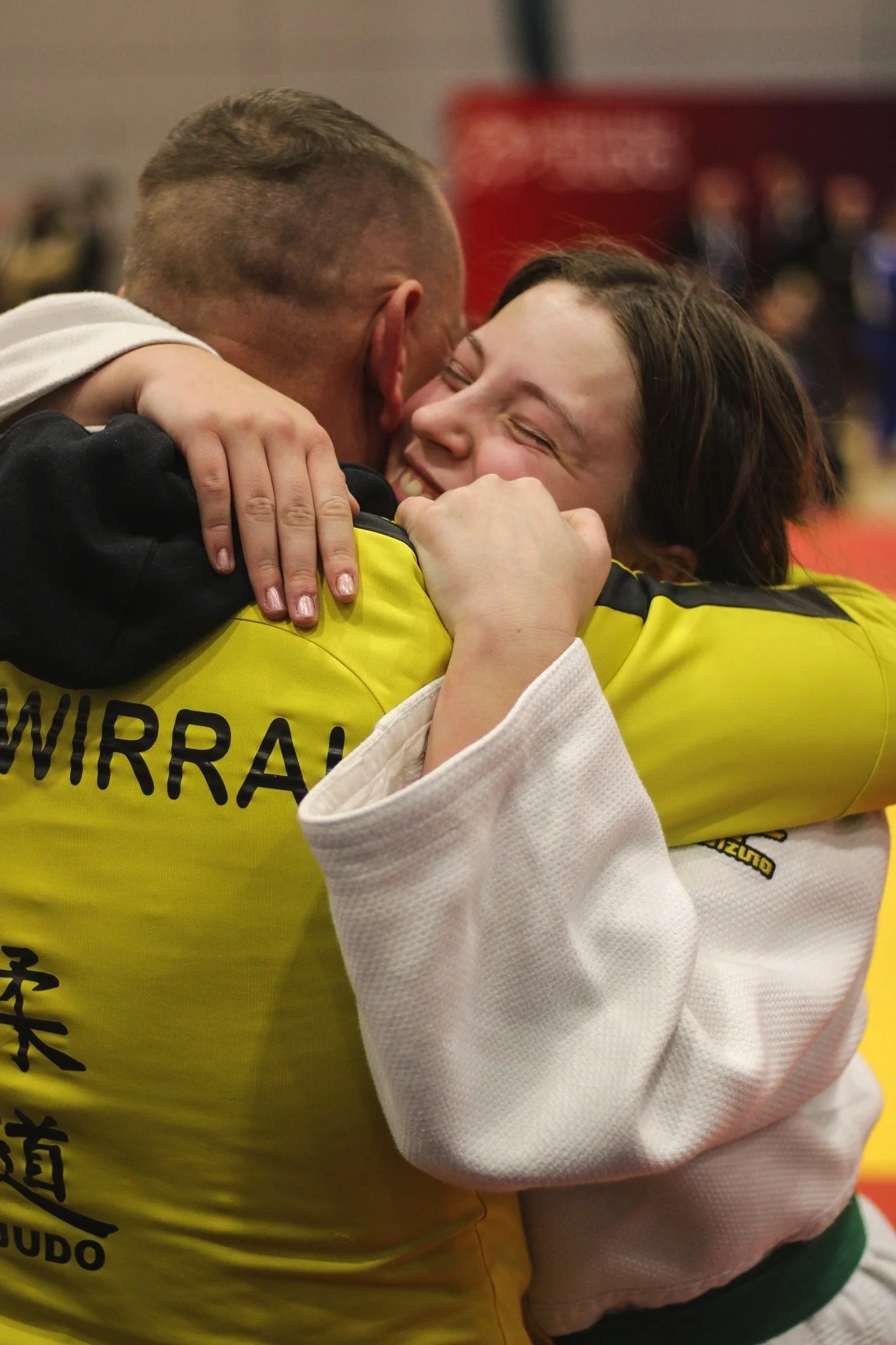 Two judo athletes embracing with happiness, one wearing a yellow judogi and the other a white judogi, in a gymnasium setting.