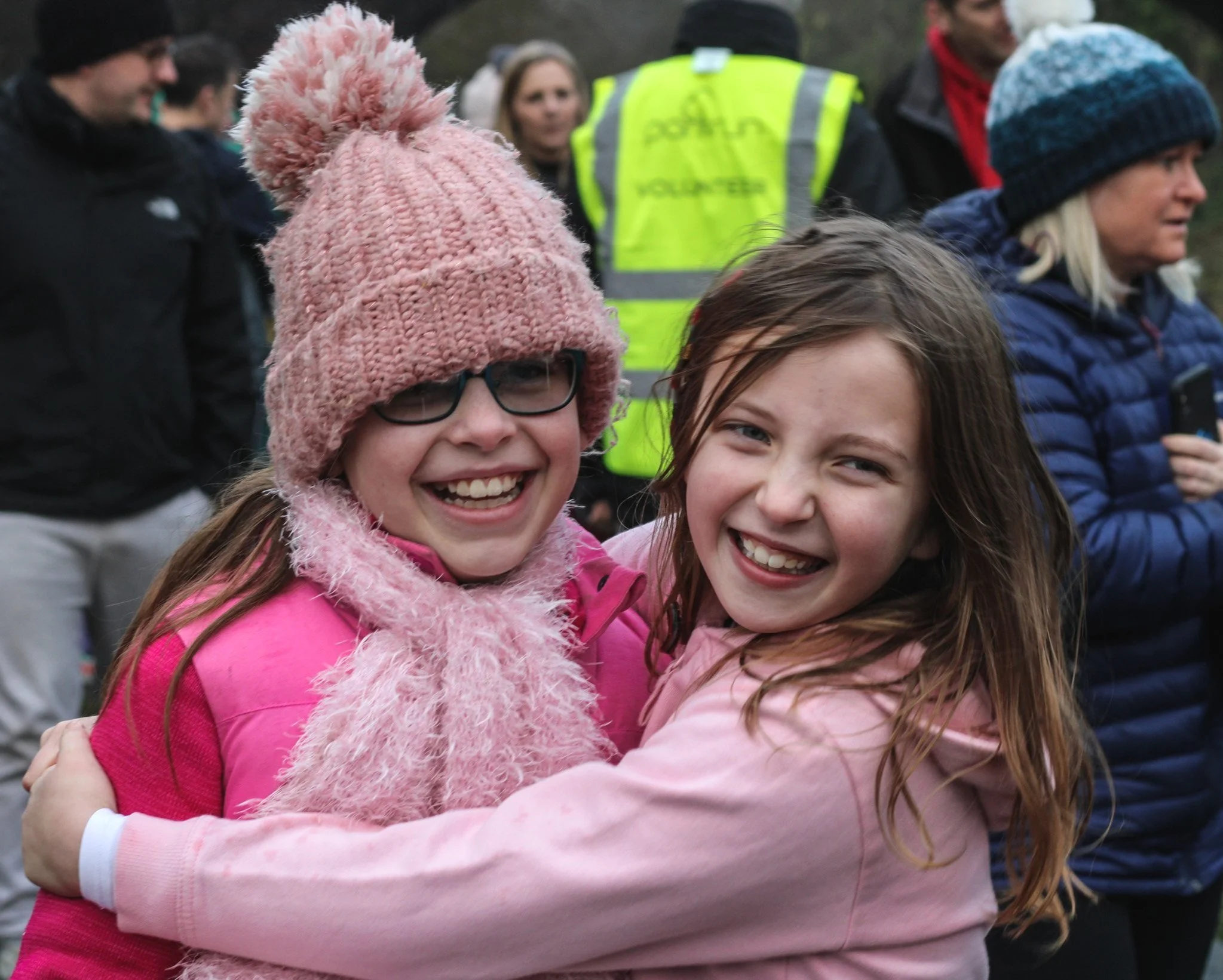 Two young girls smiling and hugging at an outdoor event, with people in jackets and a volunteer in a yellow vest in the background.