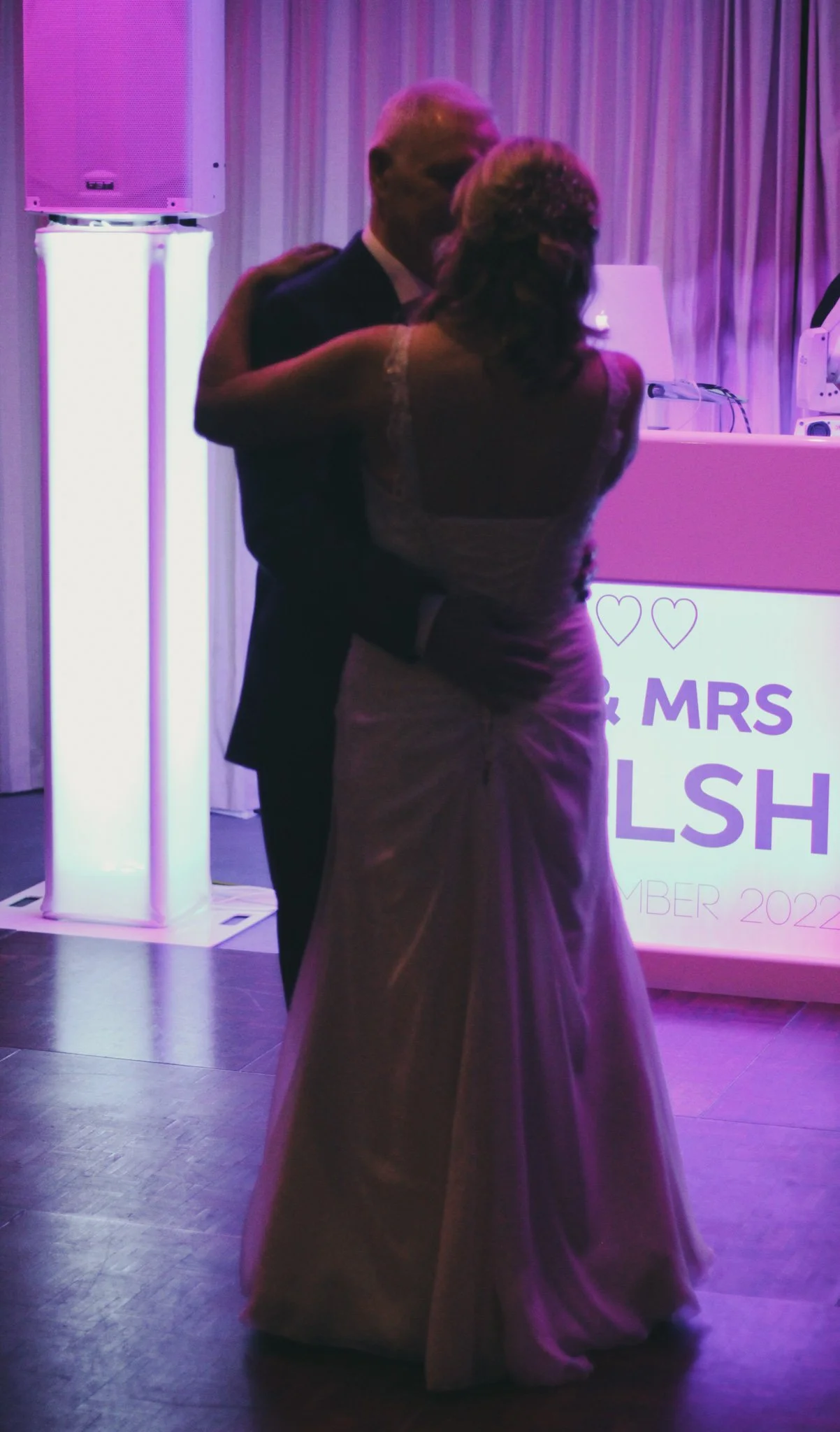 A couple in formal attire dancing at an event with purple lighting, a sign in the background reads 'MR & MRS' and '2022'.