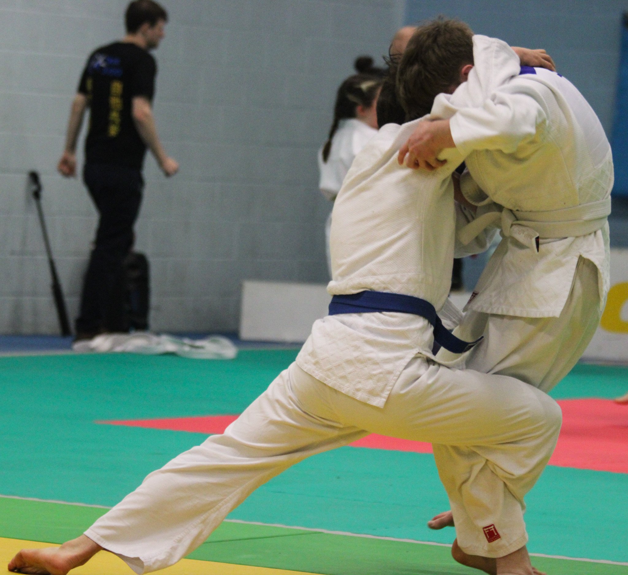 Two judo competitors in white gis engage during a match on a green and red mat, with a referee or coach and a person with a cane in the background.