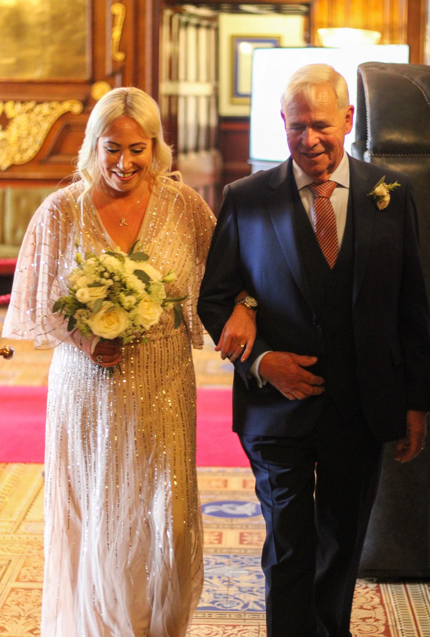 A woman in a glittery dress holding a bouquet, walking arm-in-arm with a man in a suit inside a decorated room, likely during a wedding ceremony.