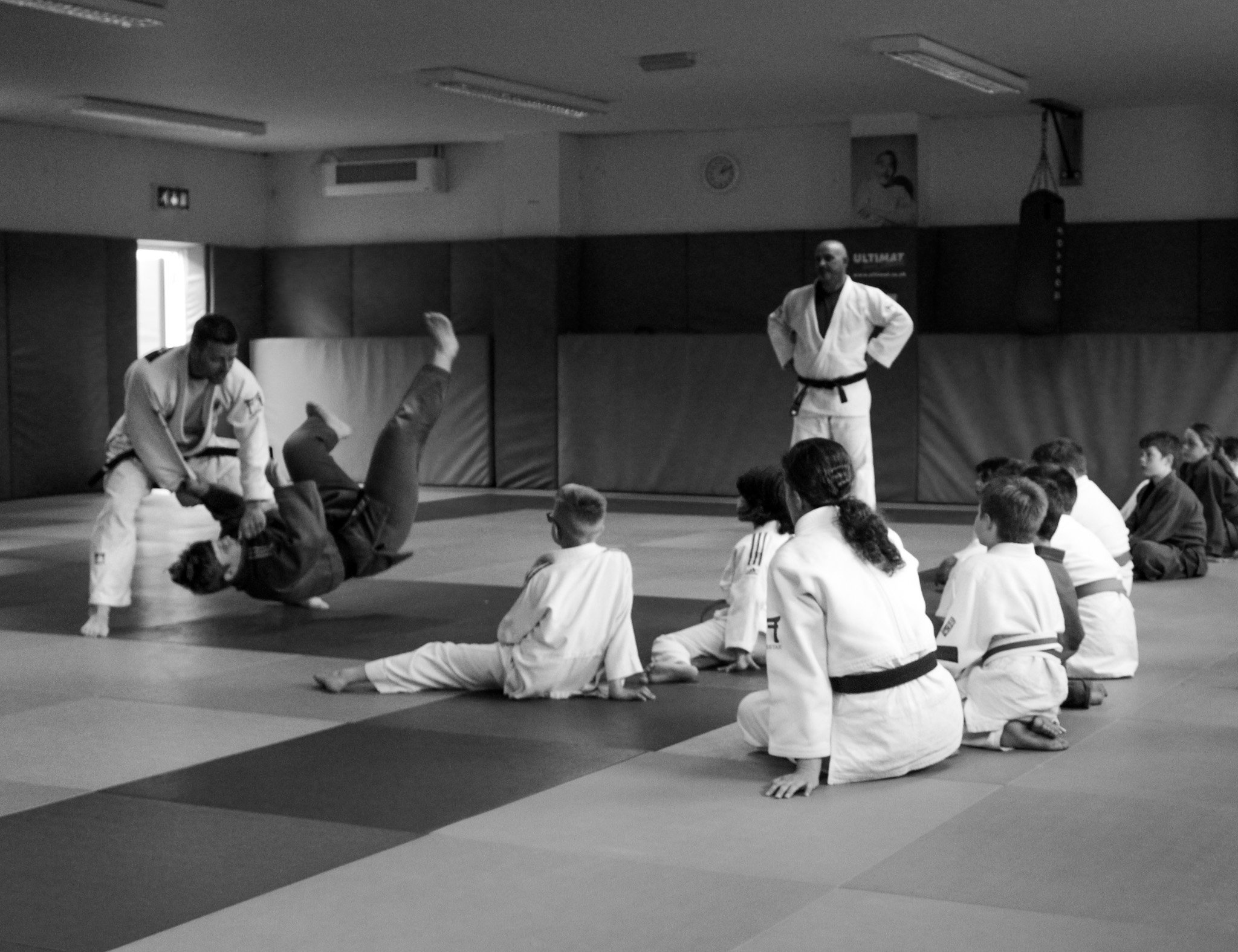 Judo class with children and two instructors practicing on mats in a dojo, black and white photo.