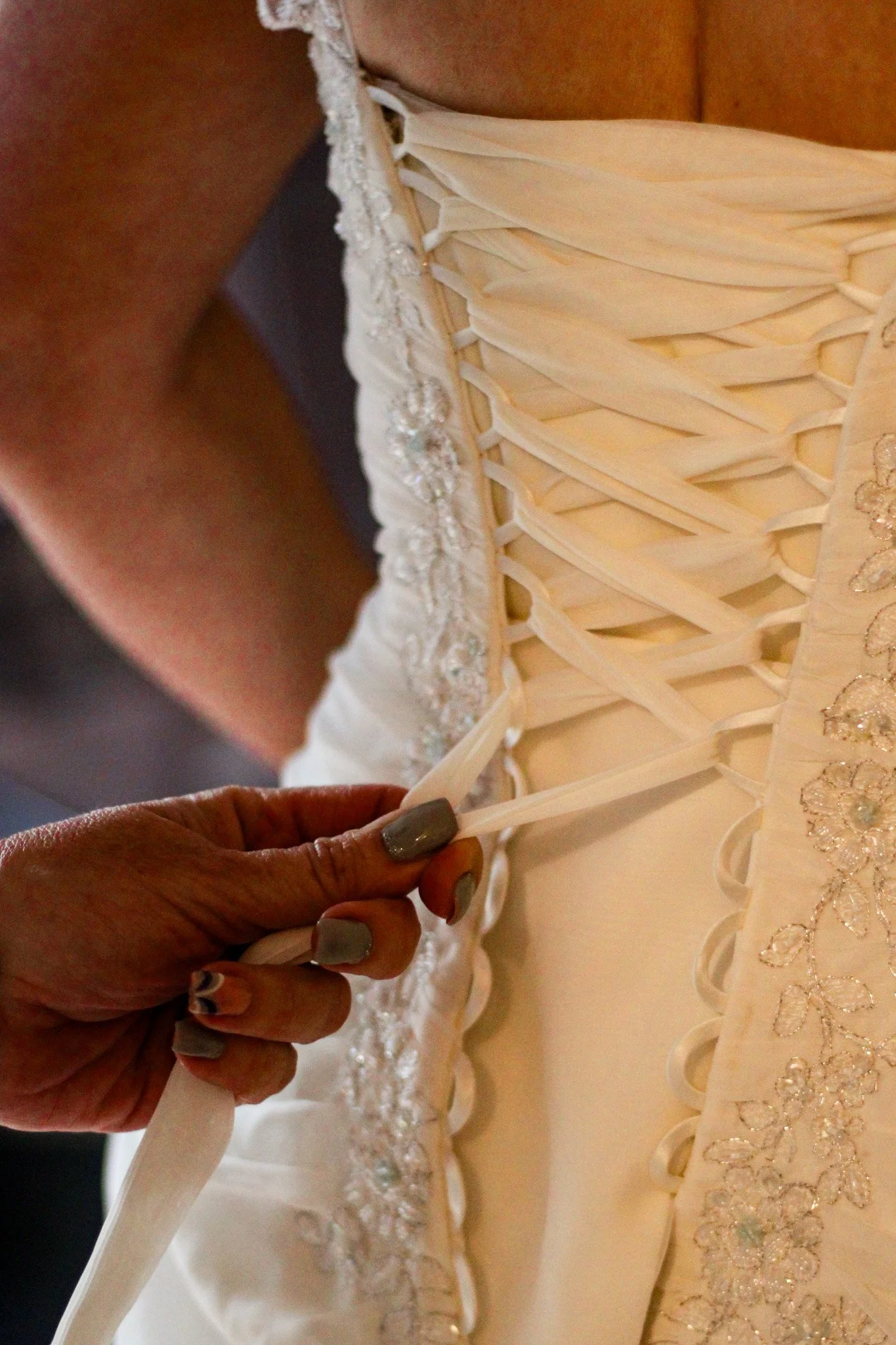 A person with painted nails fastens the laces on the back of an elegant, white wedding dress with intricate embroidery.