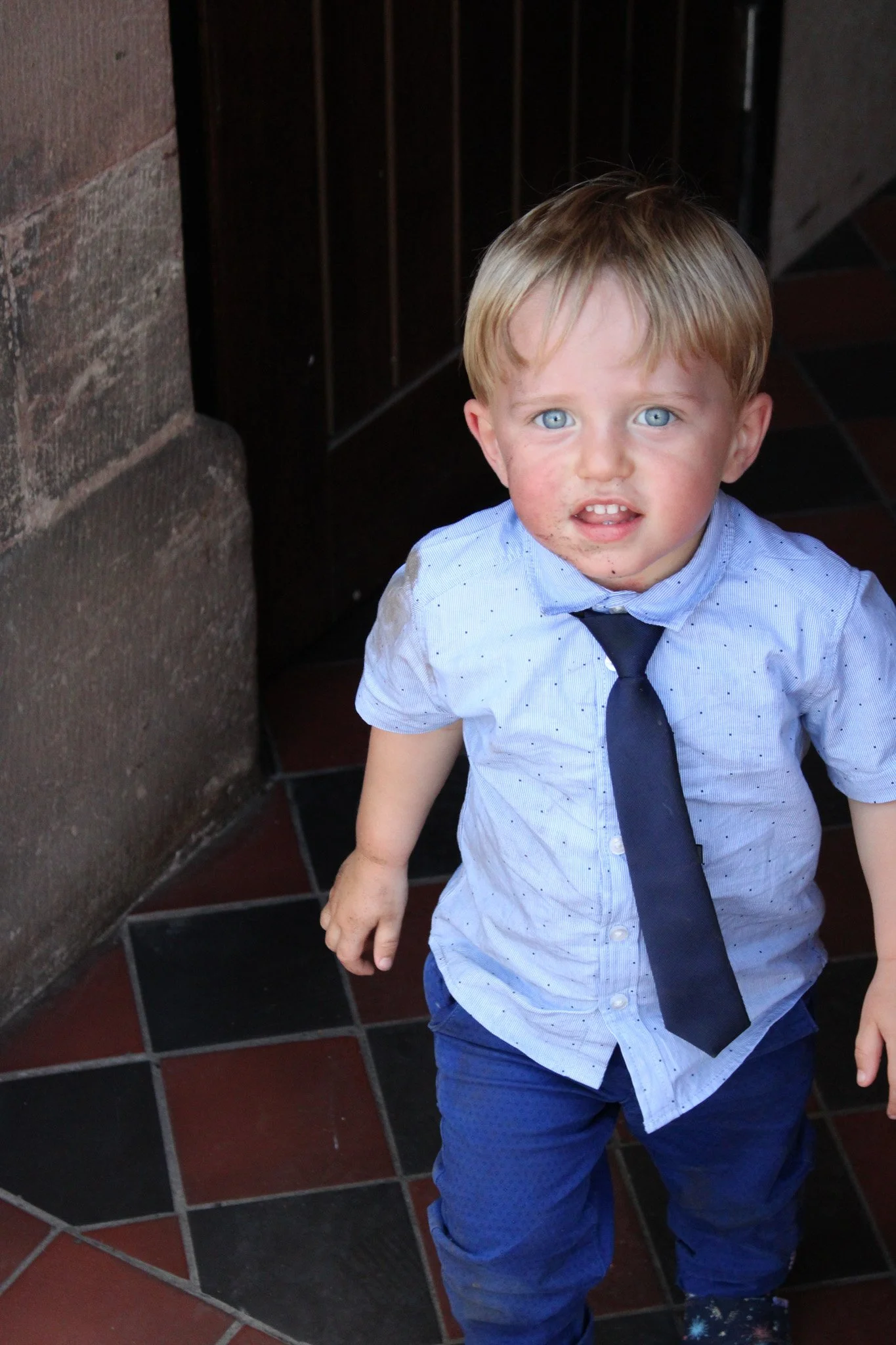 A young boy with blond hair and blue eyes, wearing a light blue shirt, dark tie, and blue pants, standing indoors with a dirty face and a surprised expression.