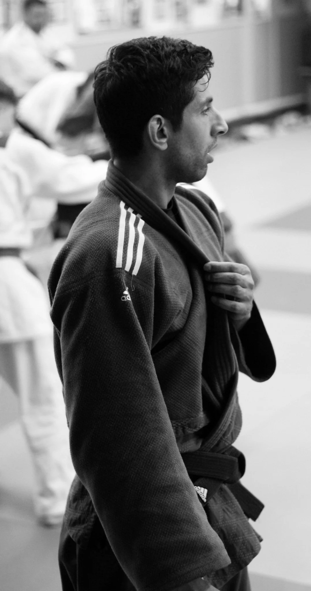 Black and white photo of a man in a judo uniform standing in a dojo, holding his jacket over his shoulder, with other people in the background.
