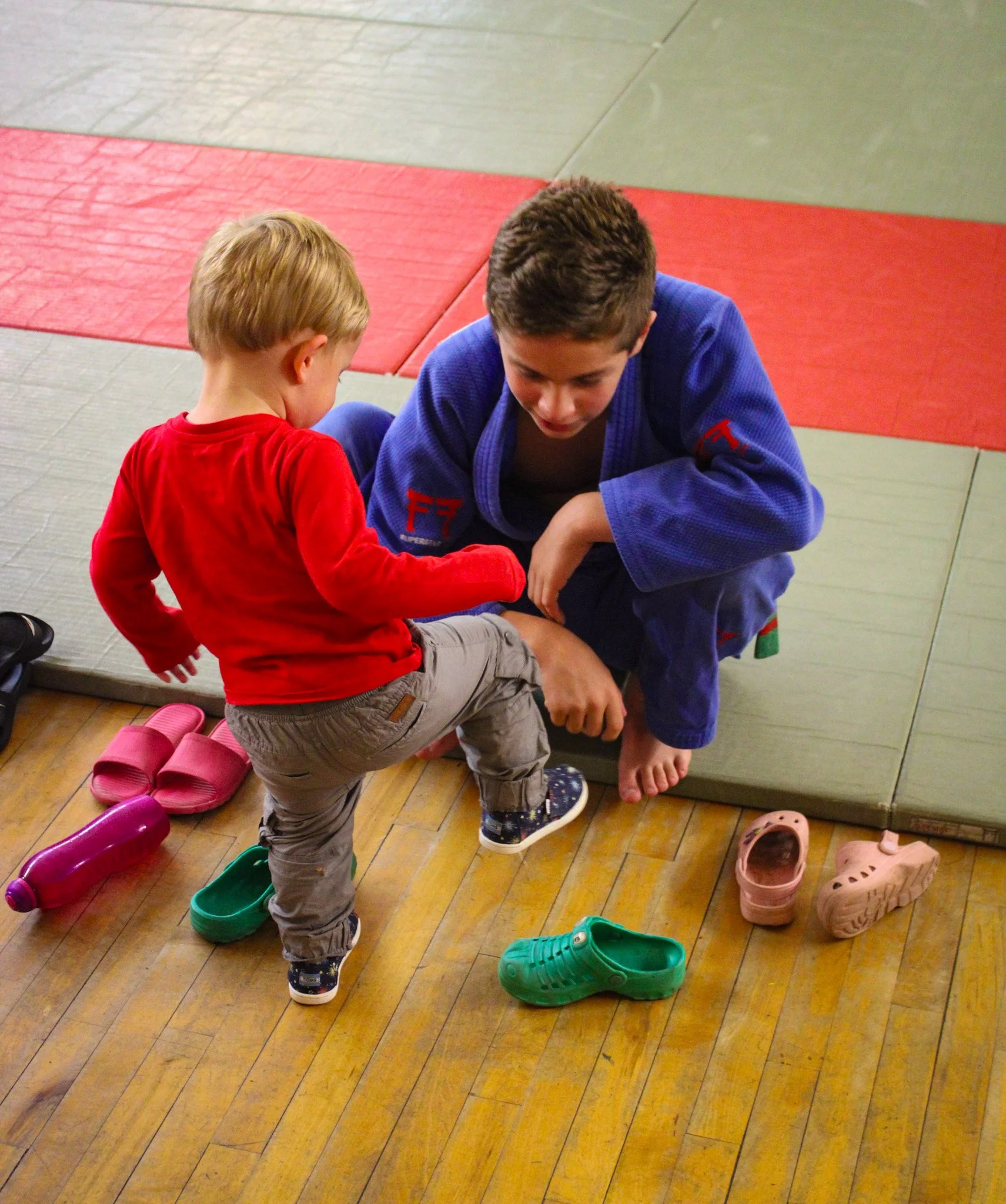 A young boy in a red shirt helps an older boy in a blue judo uniform put on or take off shoes in a gym, with several colorful shoes on the wooden floor and mats in the background.