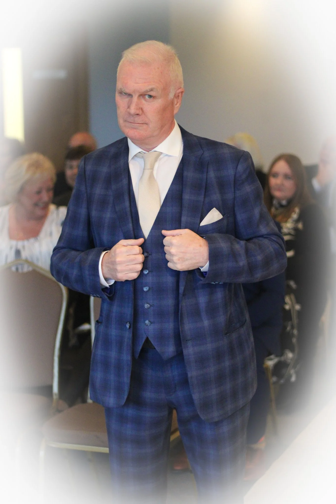 A man in a blue plaid suit with a white shirt and cream tie, adjusting his jacket during an event, with seated people in the background.