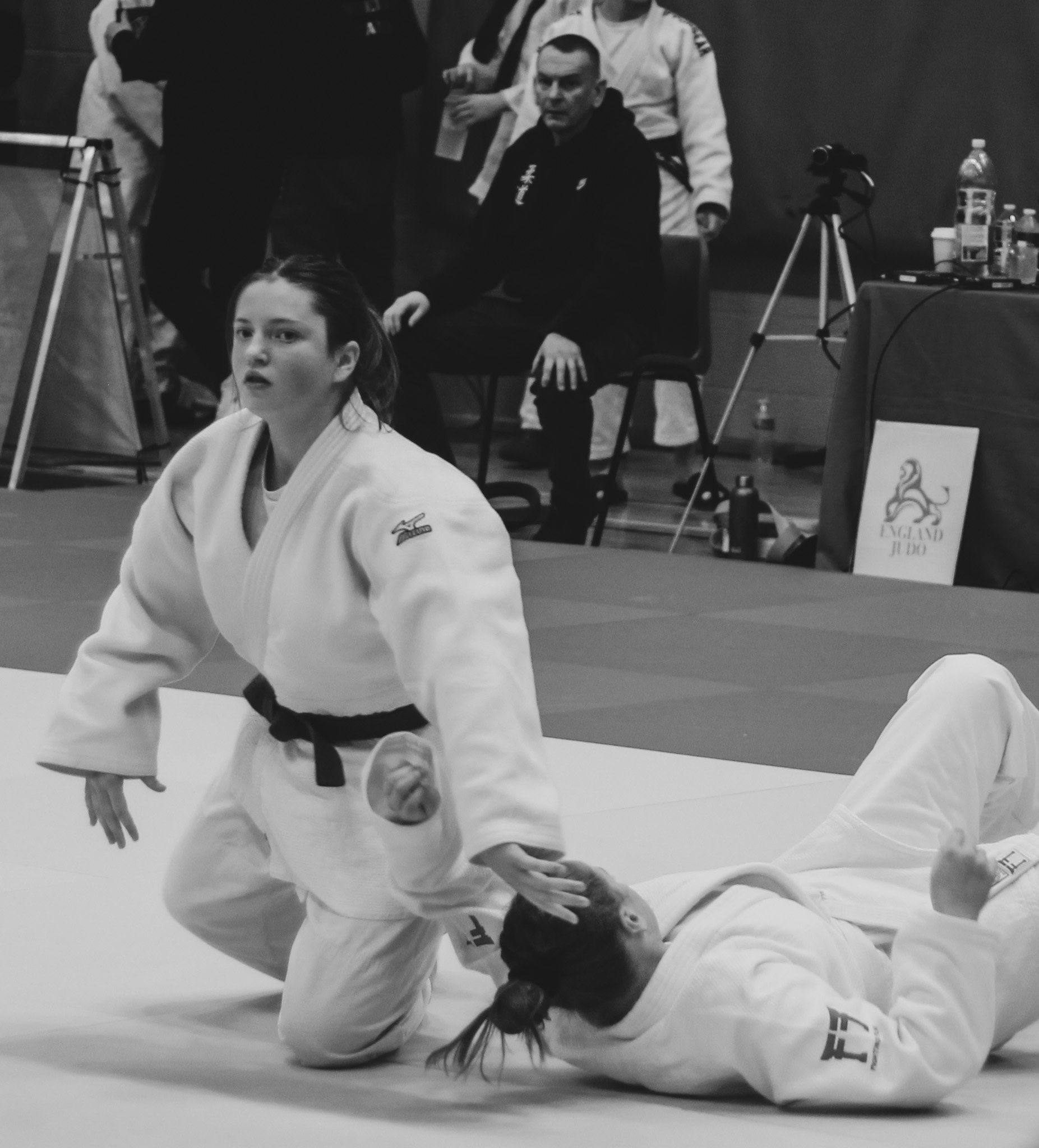 Two female judo athletes in white uniforms competing on a mat, one on top and one on the ground, with a judge seated in the background observing the match.