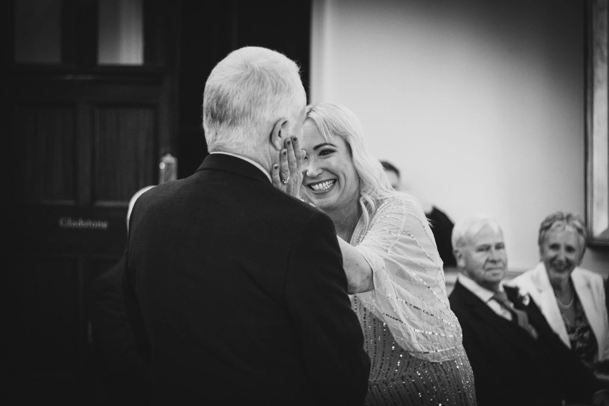 A woman with blonde hair smiling and touching the face of an older man with gray hair, as they appear to be sharing an emotional moment at a celebration or gathering, with two other older individuals seated in the background.