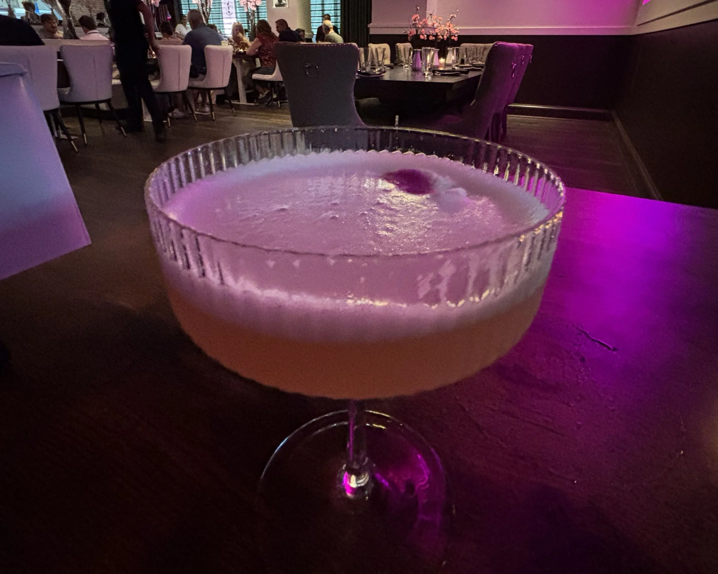 A cocktail in a stemmed glass with a frothy top, placed on a dark wooden table in a dimly lit restaurant with purple pink lighting, with other diners in the background.