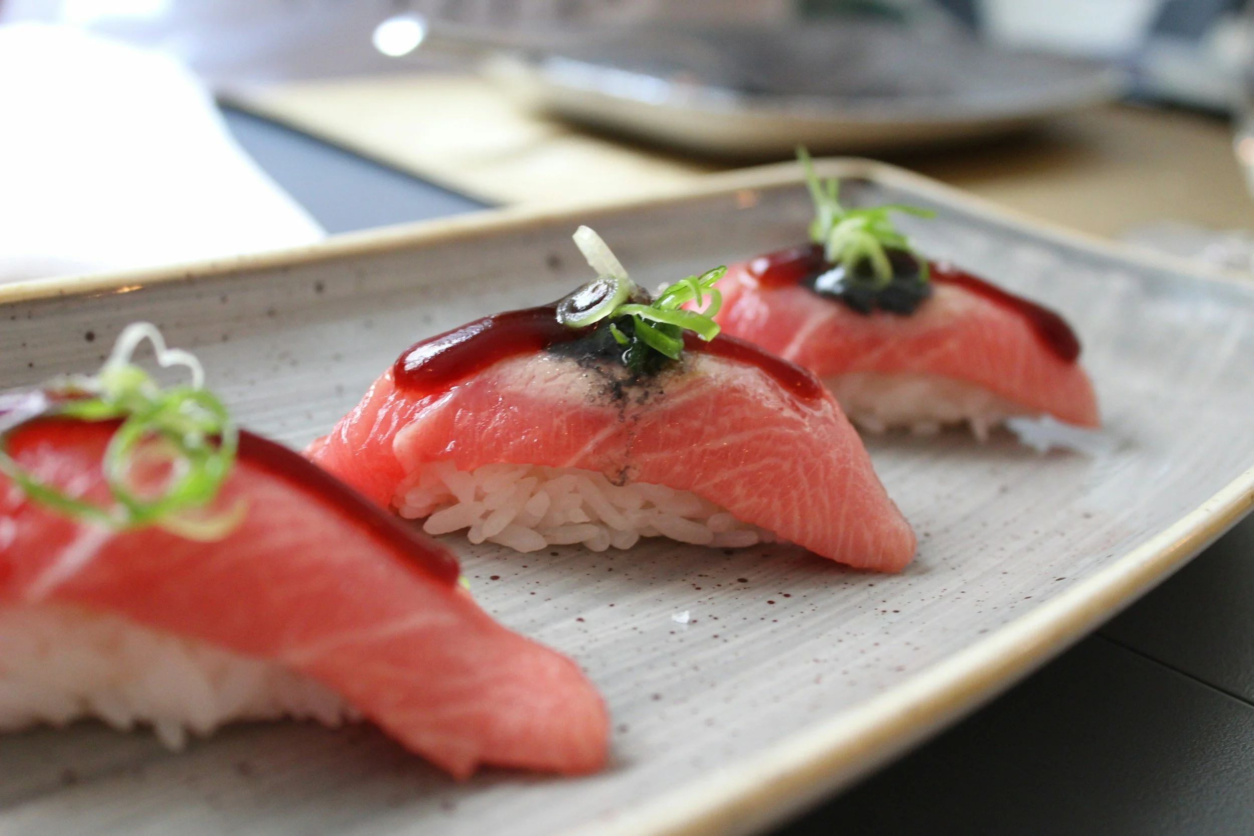 Three pieces of sushi with sliced raw fish topped with green onions and dark sauce, arranged on a rectangular ceramic plate.