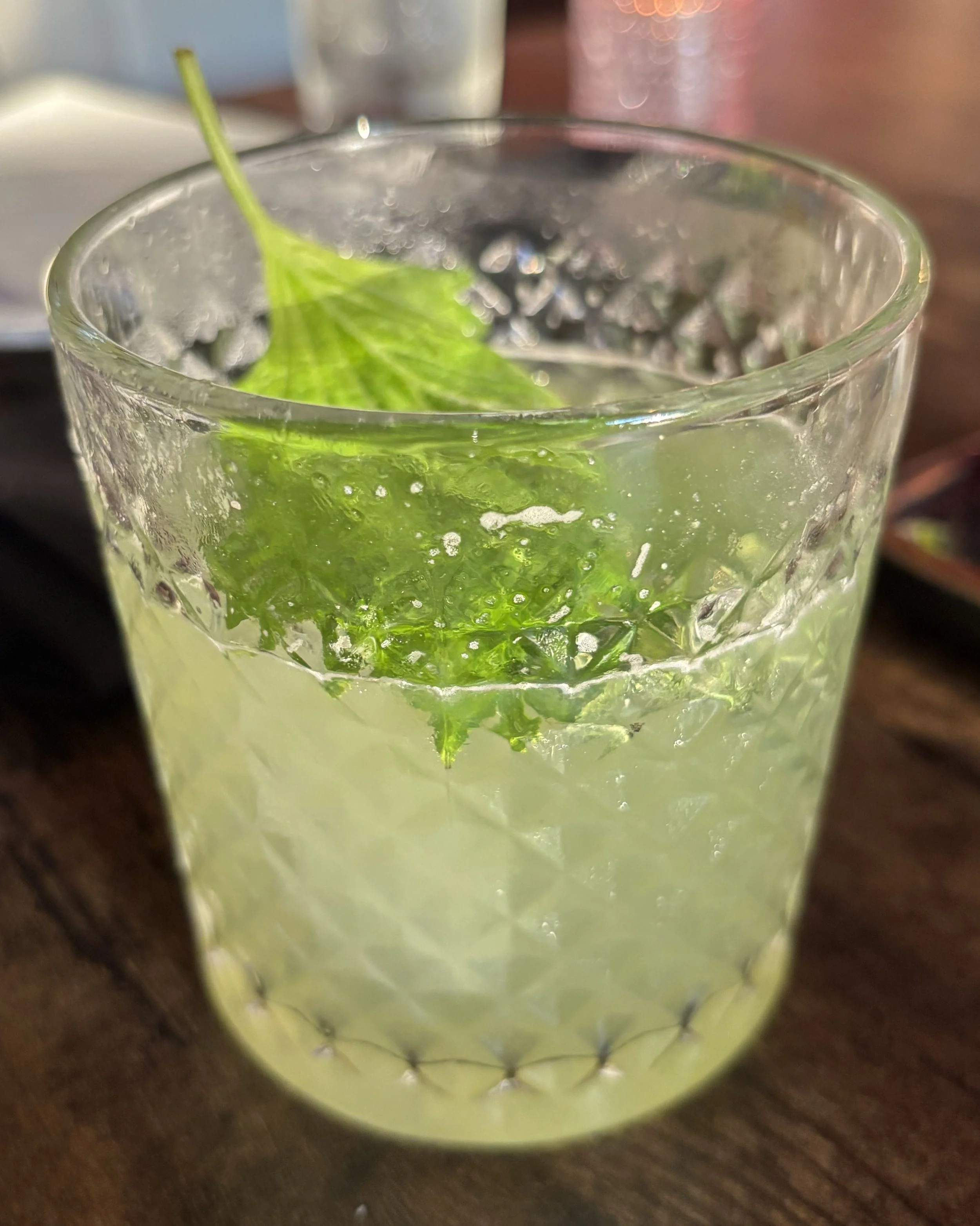 A textured glass tumbler contains a lime and mint beverage with a mint leaf on top, placed on a wooden surface.