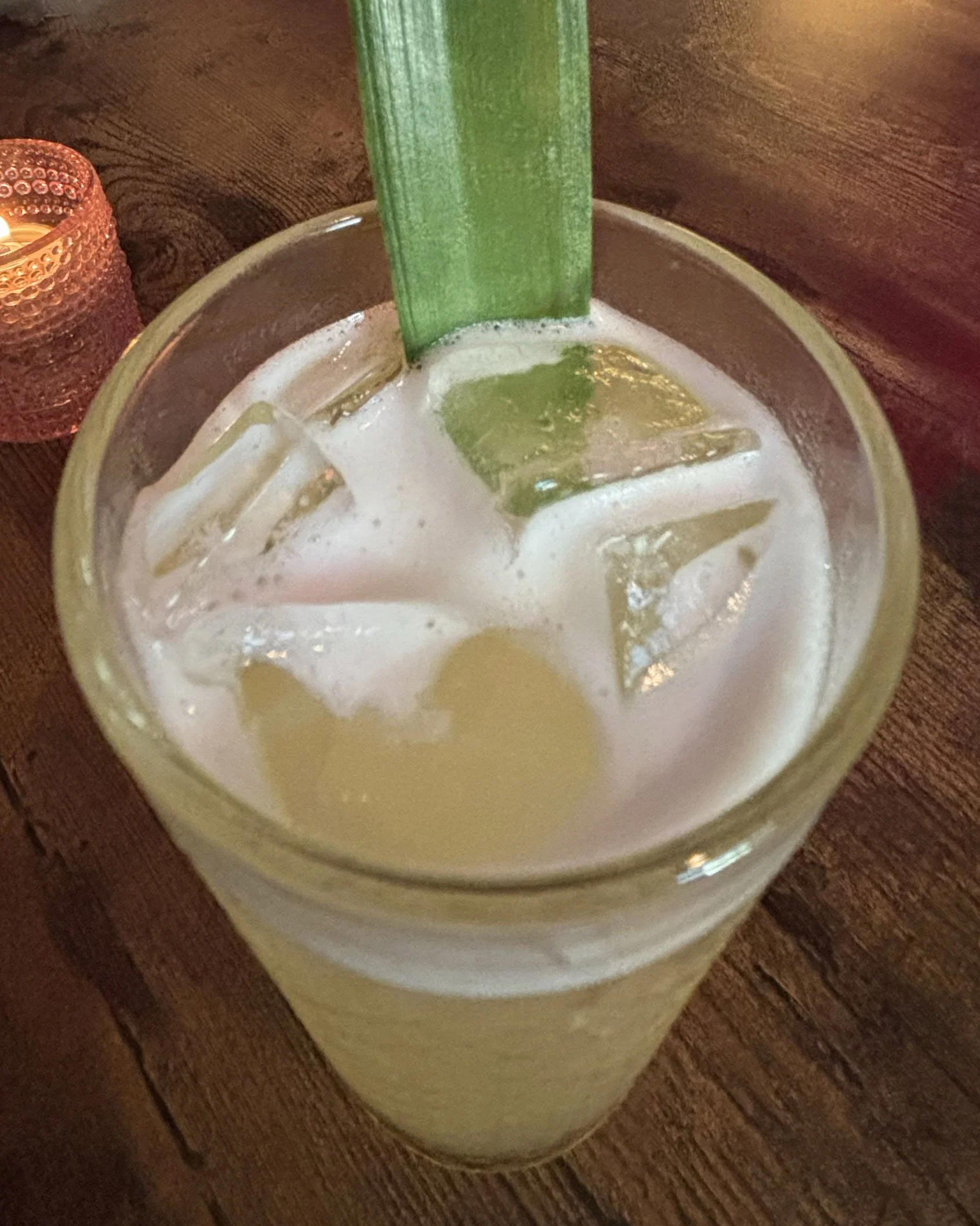 A glass of lemonade with ice cubes and a green straw on a wooden table.