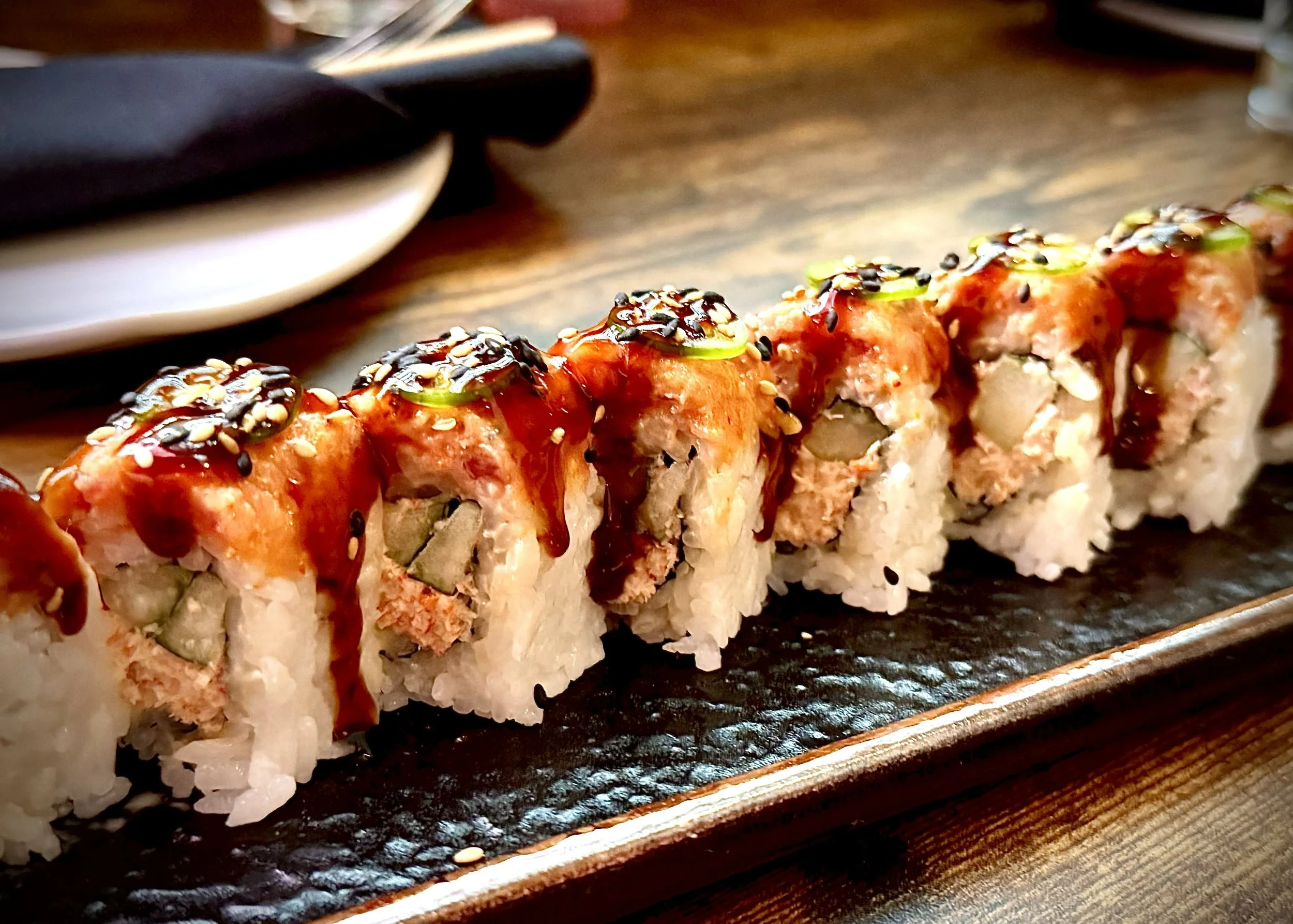 Close-up of a sushi roll topped with sauce, green onion, and black sesame seeds, served on a rectangular black plate.