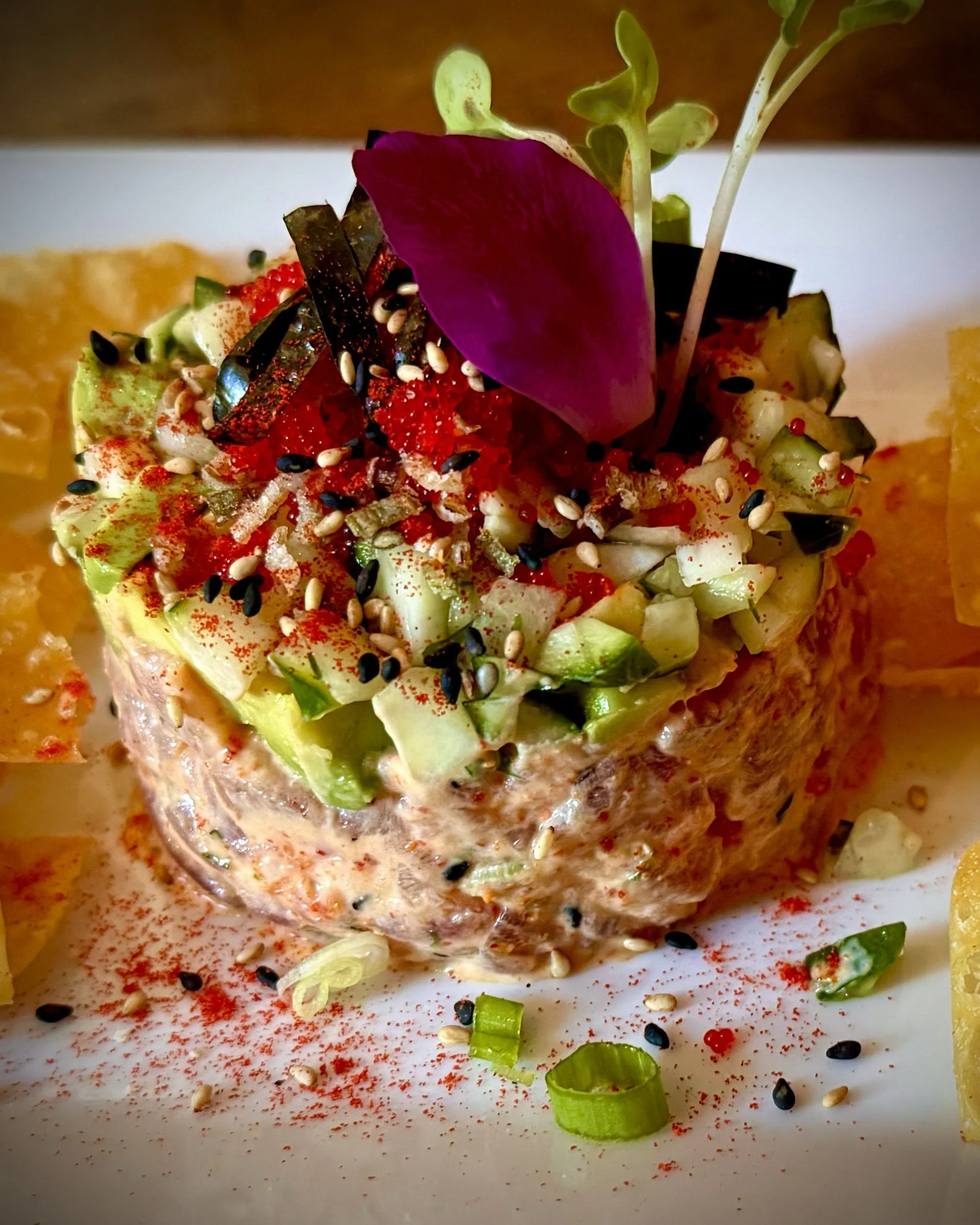 A close-up of an appetizer featuring a cylindrical salmon mixture topped with diced avocado, red caviar, sesame seeds, and garnished with purple and green herbs, on a white plate with lemon wedges and a sprinkle of paprika.