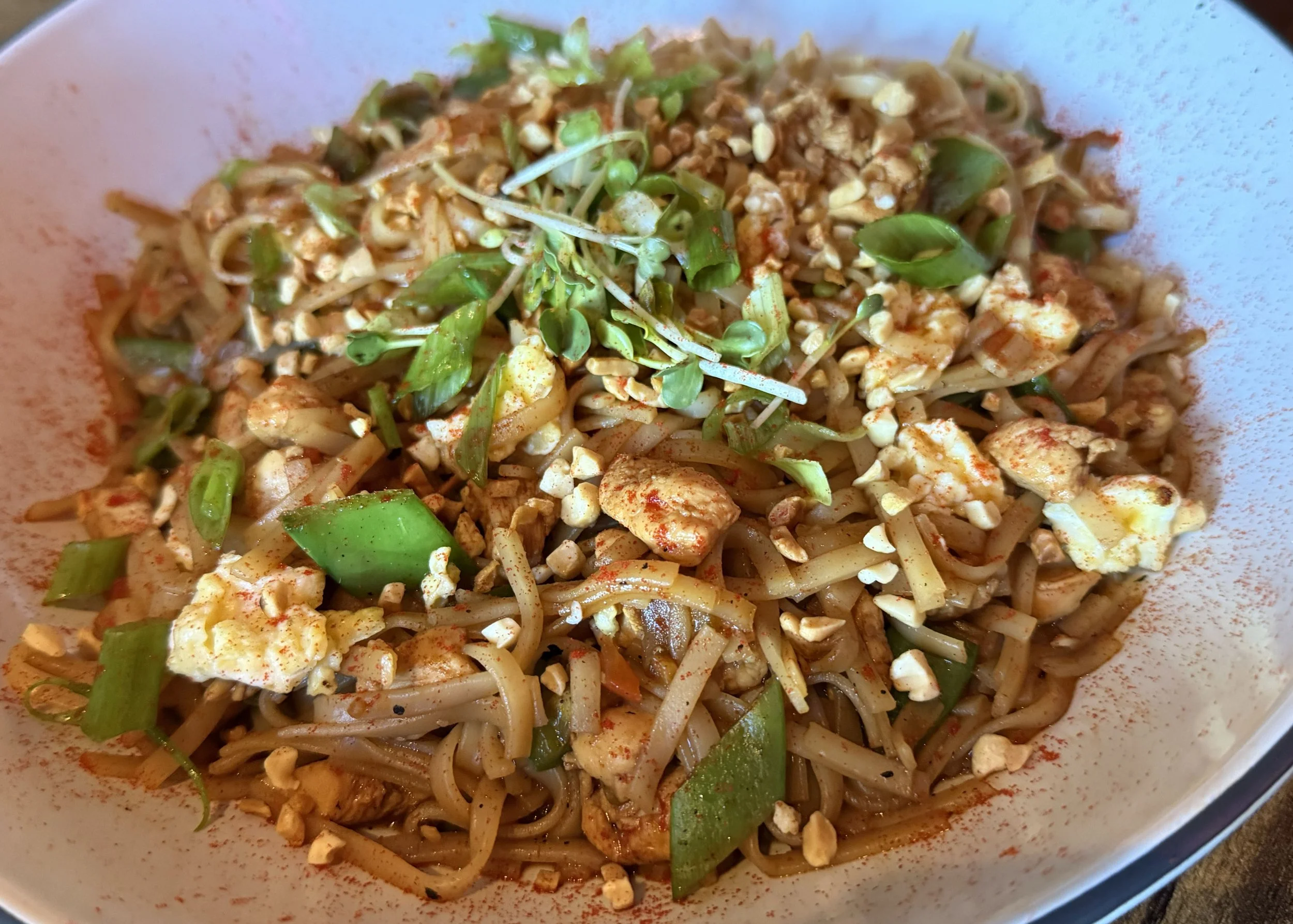 Close-up of a bowl of Asian-style noodle stir-fry with chopped vegetables, scrambled egg pieces, crushed peanuts, and green garnishes.