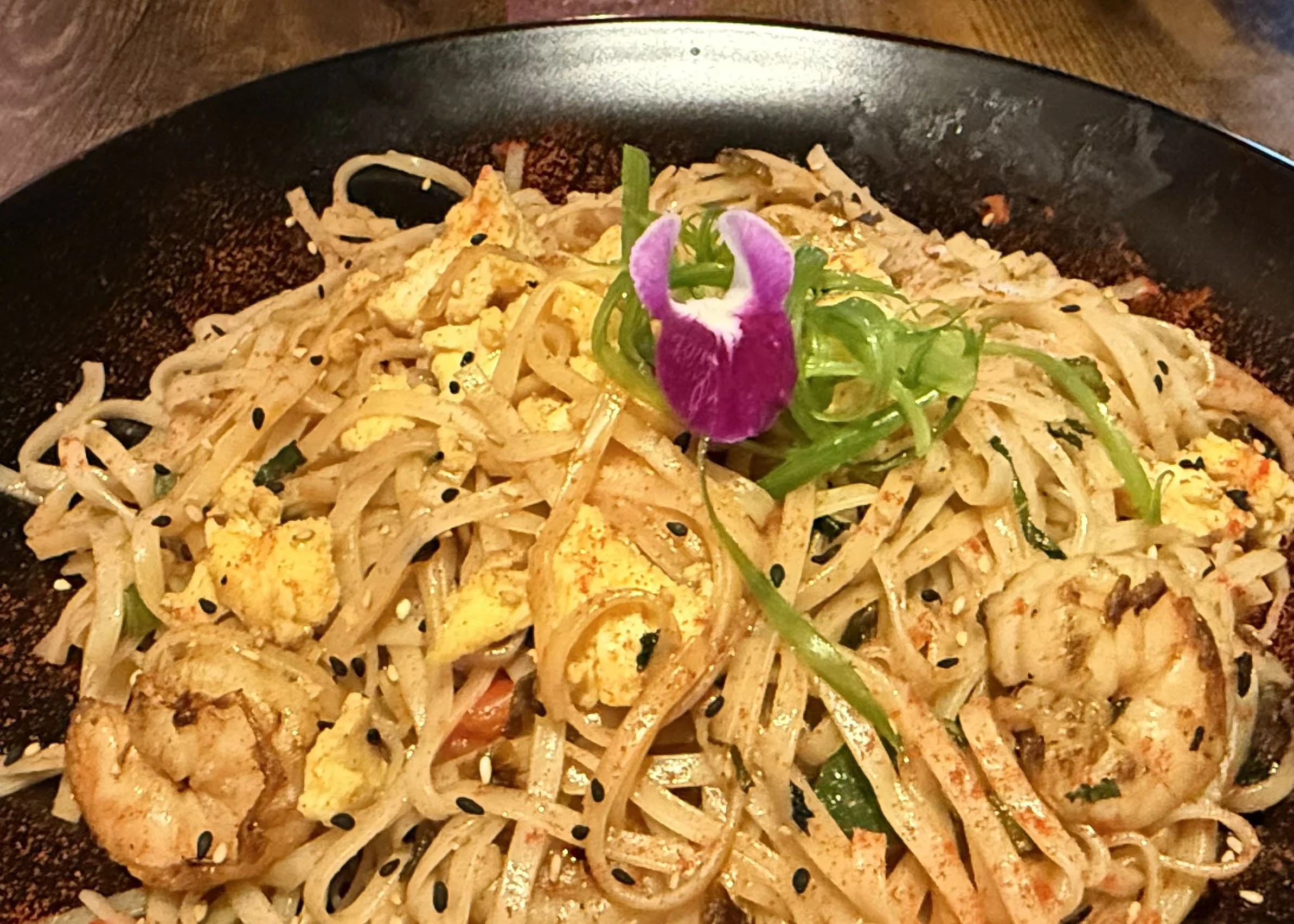 A plate of Asian-style noodle stir-fry garnished with a purple and white flower.