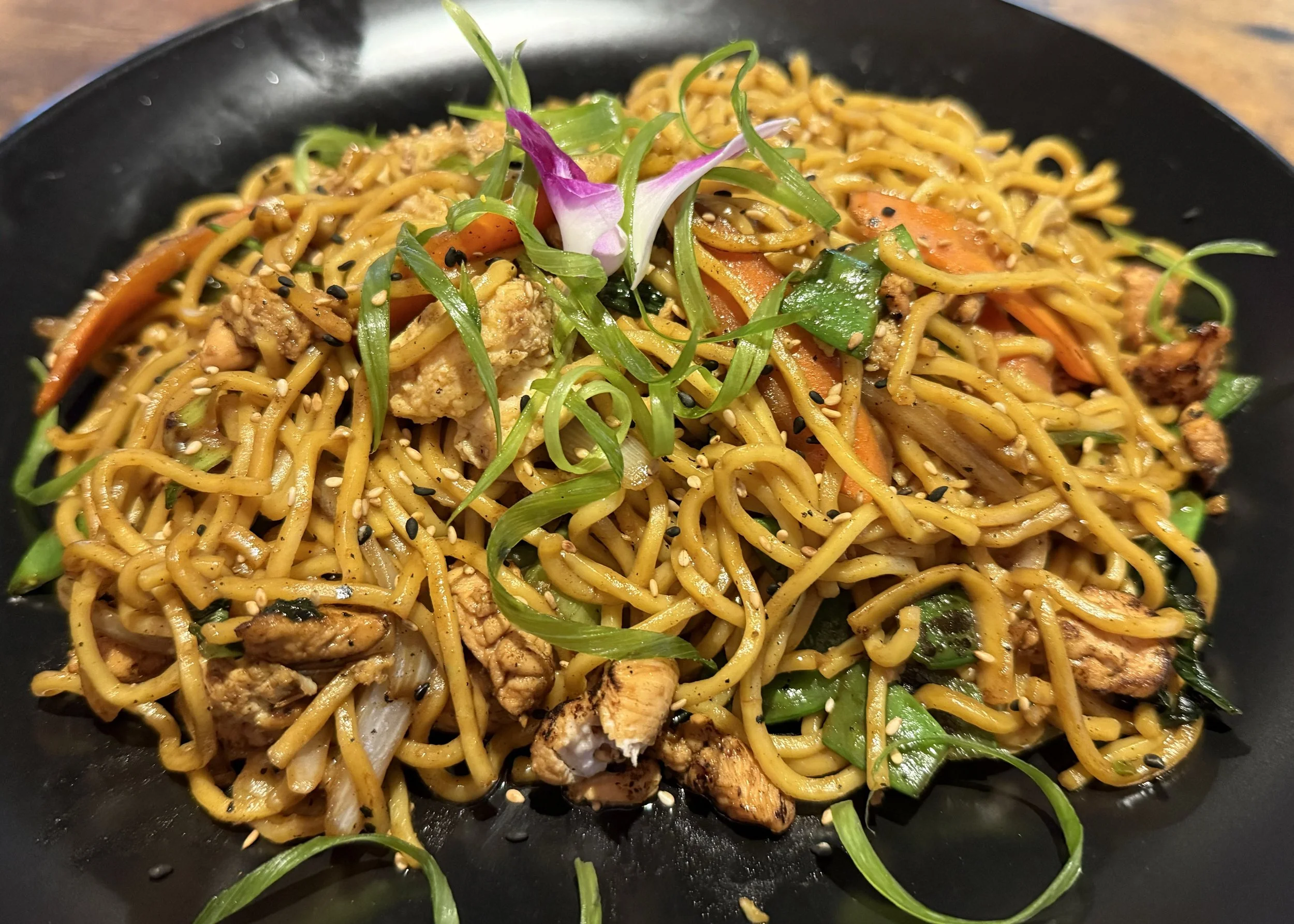 A black plate of stir-fried noodles with vegetables and chicken, garnished with green onions and sesame seeds.