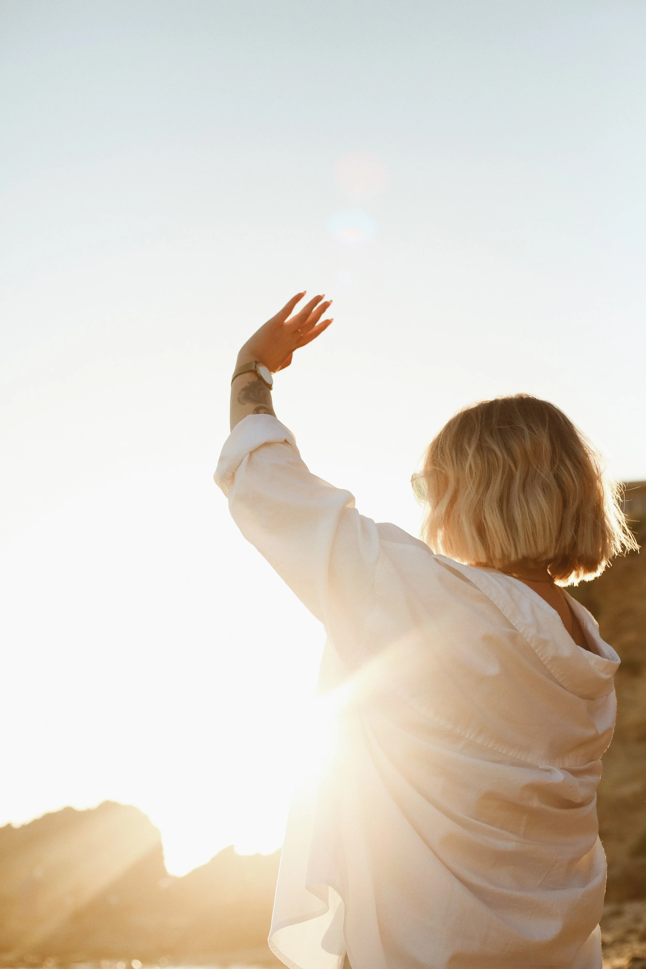 Person with blond hair wearing a white jacket reaching towards the sky during sunset.