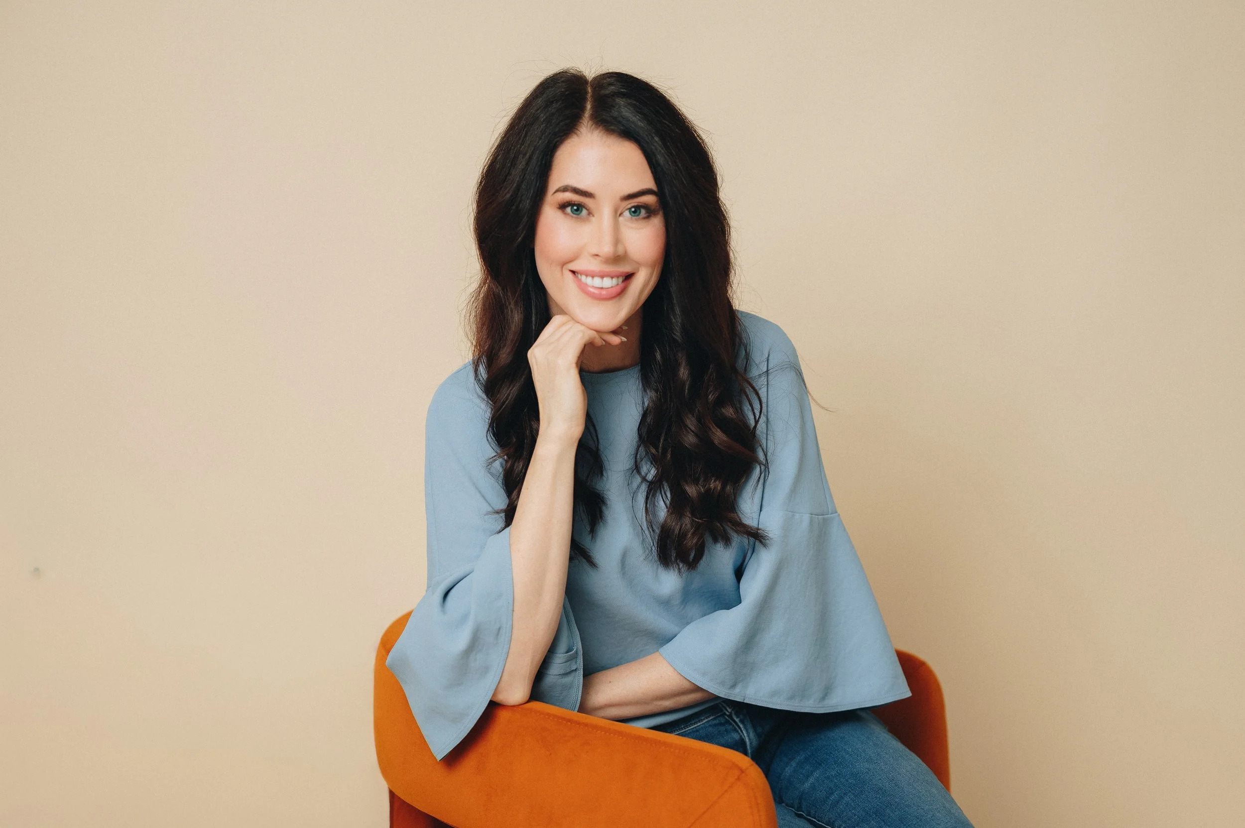 A woman with long dark wavy hair, wearing a light blue blouse and jeans, smiling and resting her chin on her hand while sitting in an orange chair against a plain beige wall.