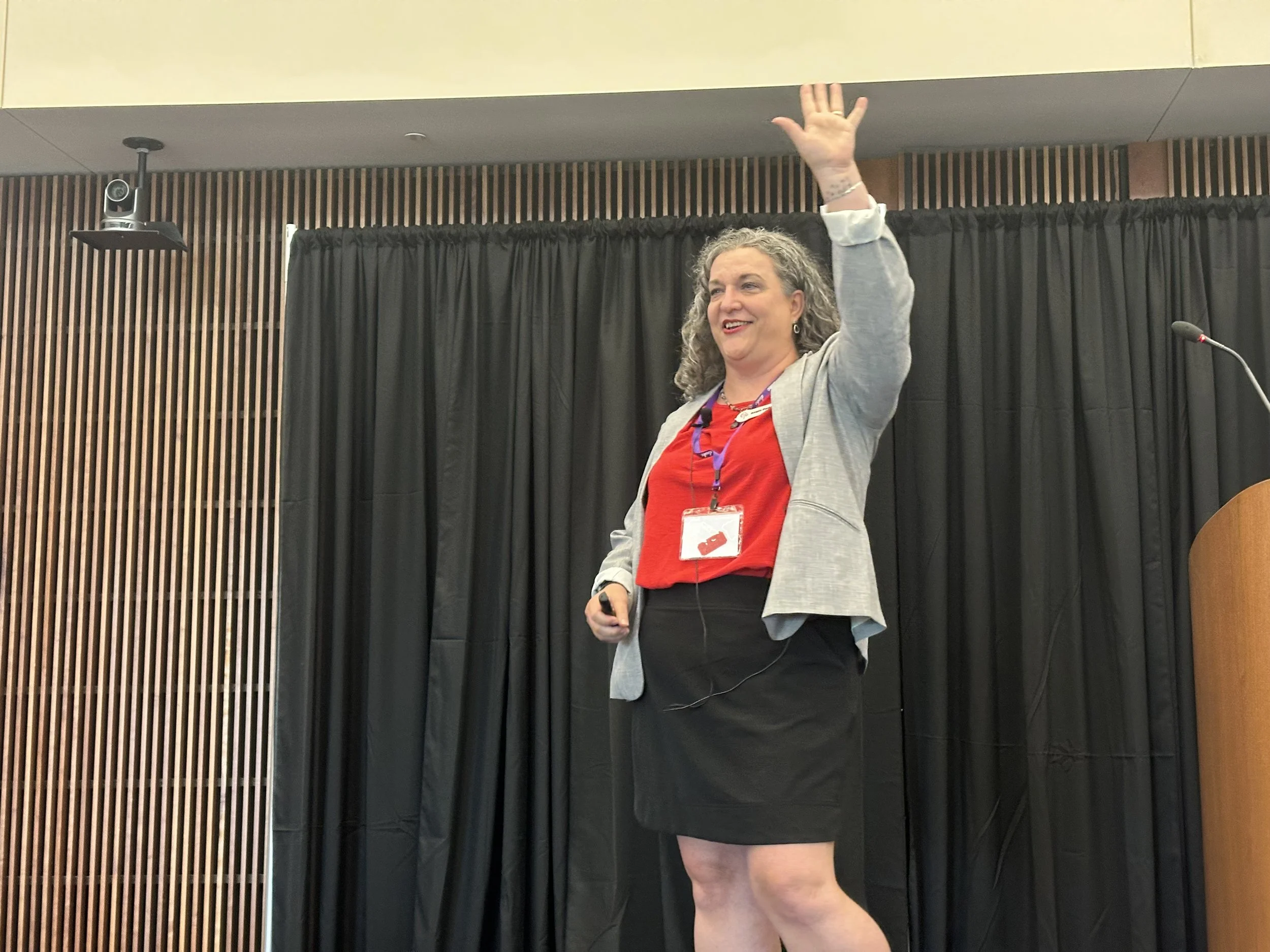 A woman with curly gray hair is giving a presentation or speech on a stage, waving to the audience with her right hand, wearing a red shirt, gray blazer, and black skirt, with a lanyard and badge around her neck.
