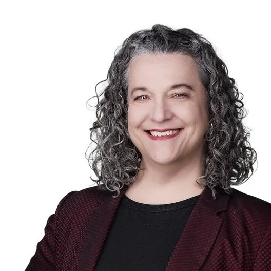 A caucasian woman with shoulder-length curly brown and gray hair is smiling. She is wearing a dark red textured blazer over a black top, in front of a plain white background.