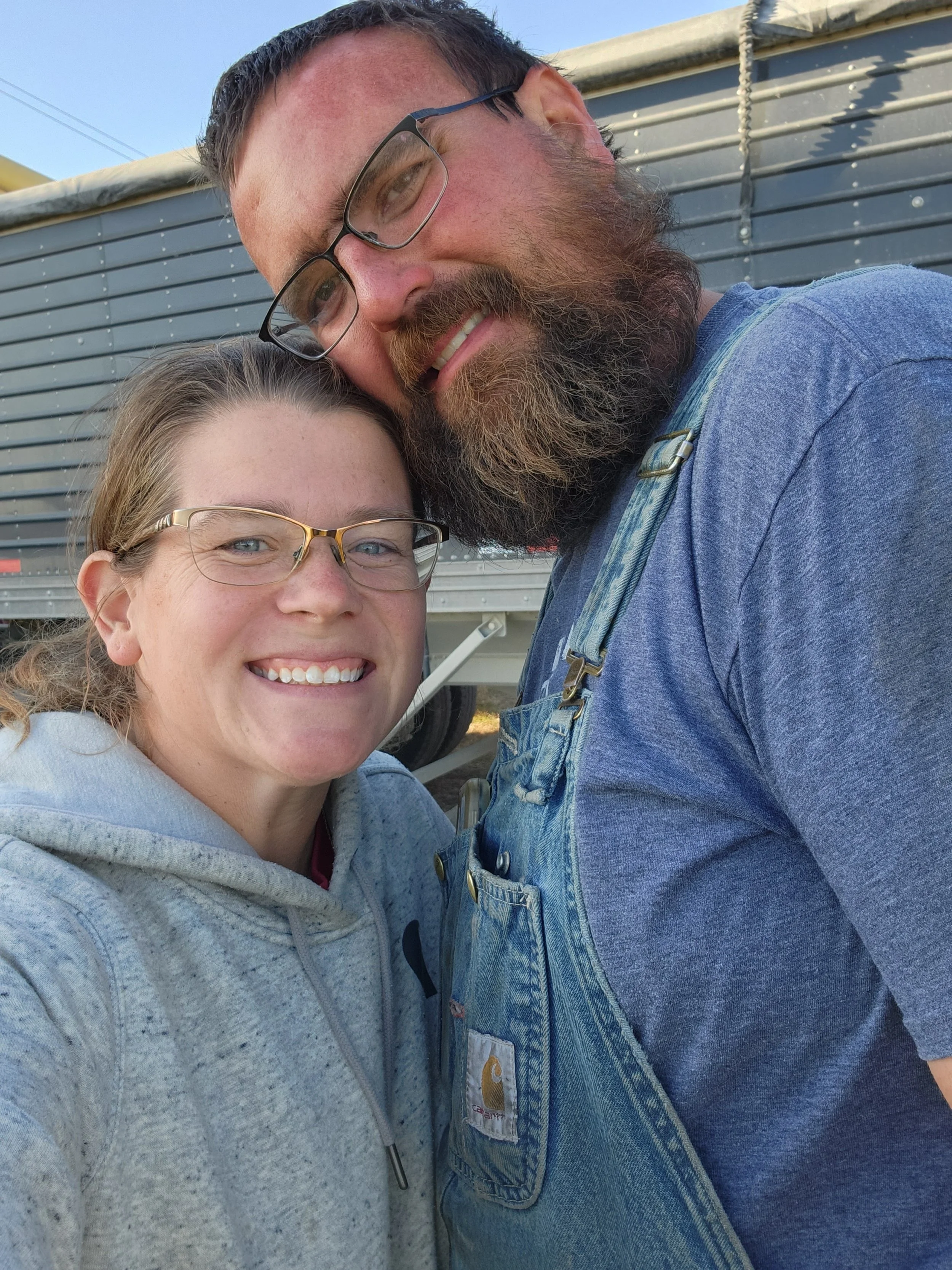 Two people smiling, one wearing glasses and a gray hoodie, the other with a beard, glasses, and denim overalls, standing close together in front of a large vehicle or equipment.