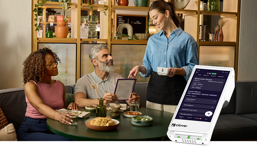 A waitress takes an order from a woman and man seated at a restaurant table with food and drinks. A handheld device displays order details.