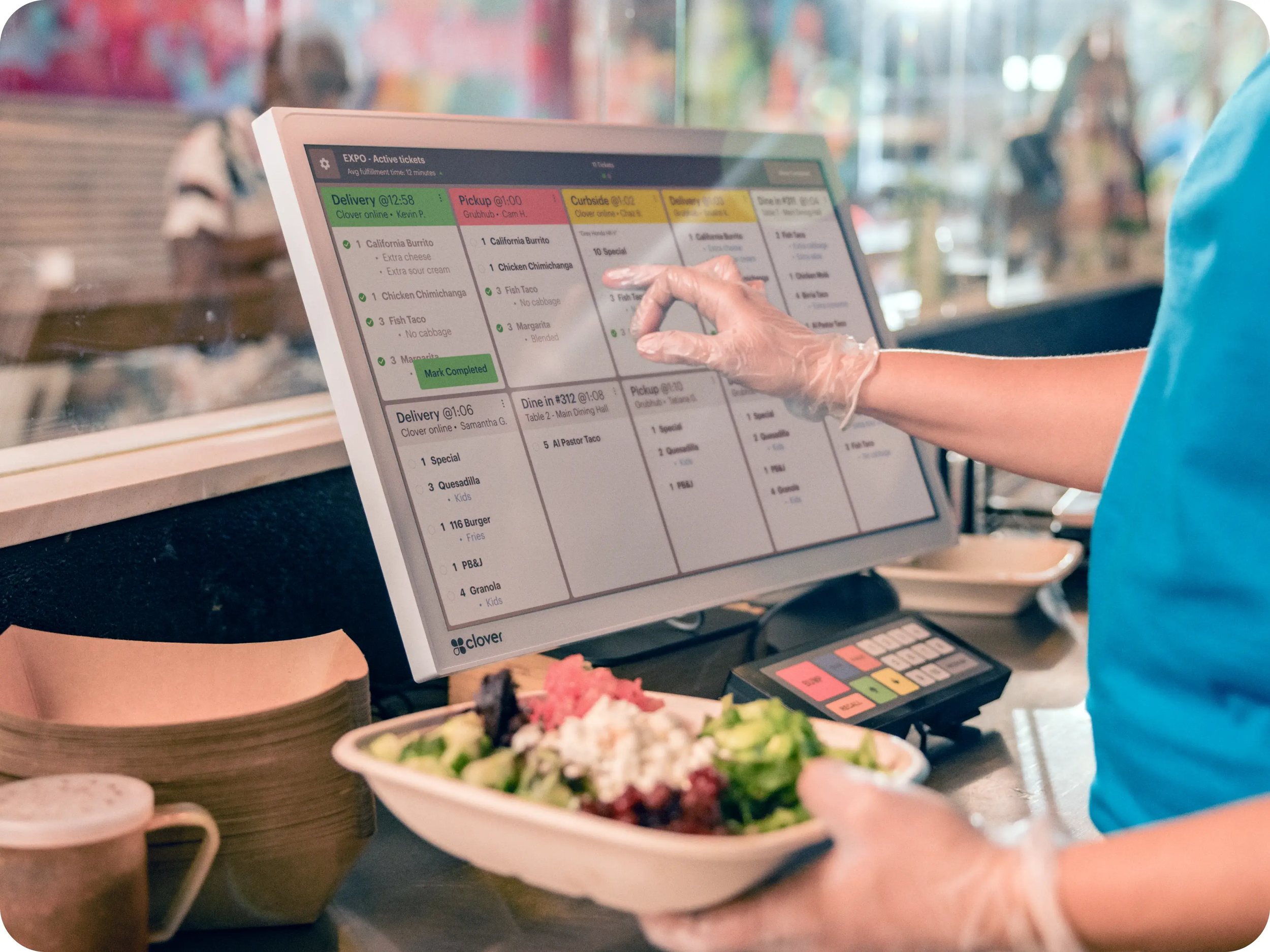 A person in a blue shirt using a touchscreen ordering kiosk at a restaurant, with a bowl of salad and paper containers on the counter.