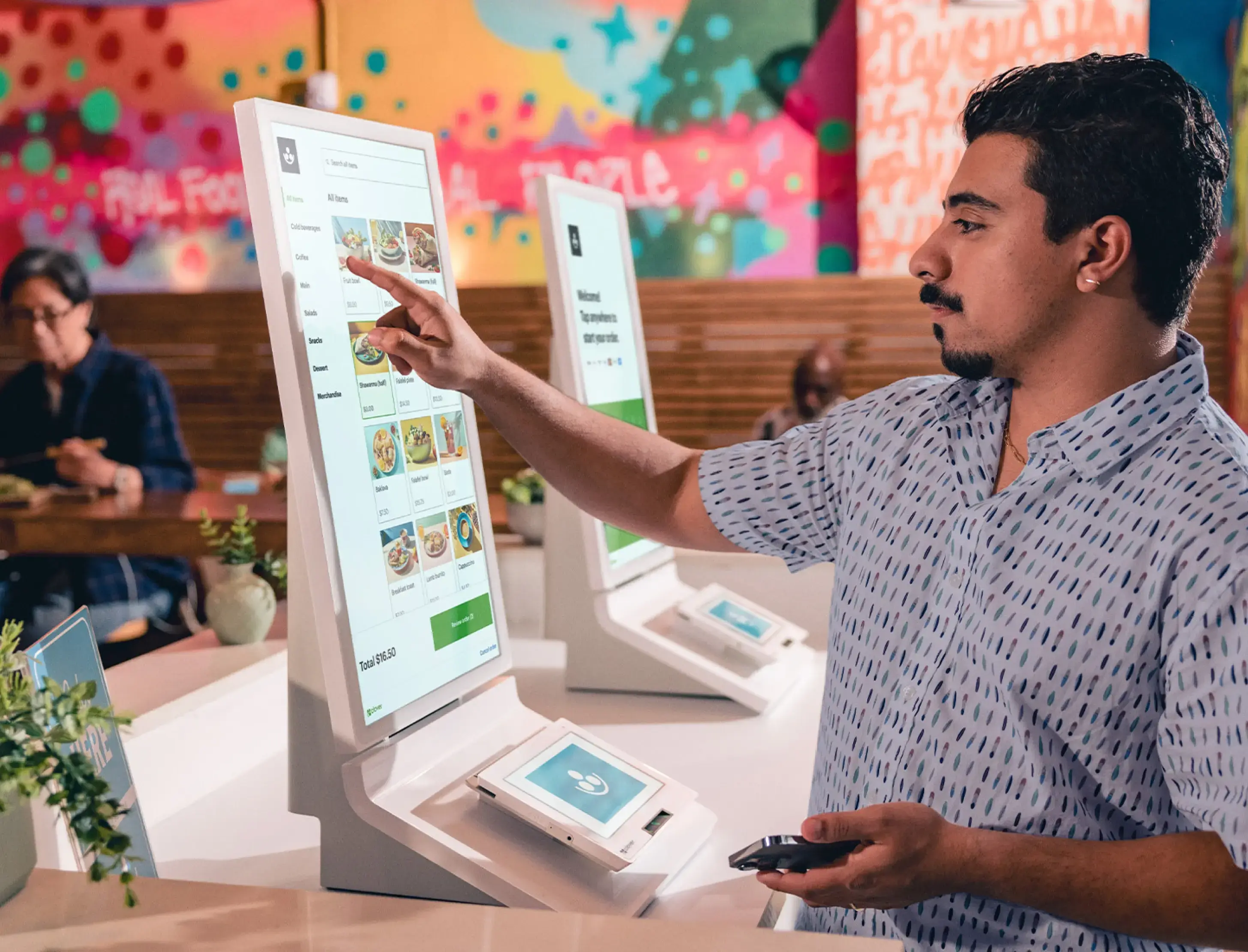 A man in a patterned shirt uses a touchscreen ordering kiosk at a restaurant. Other people are seated at tables in the background, and colorful wall art decorates the background.