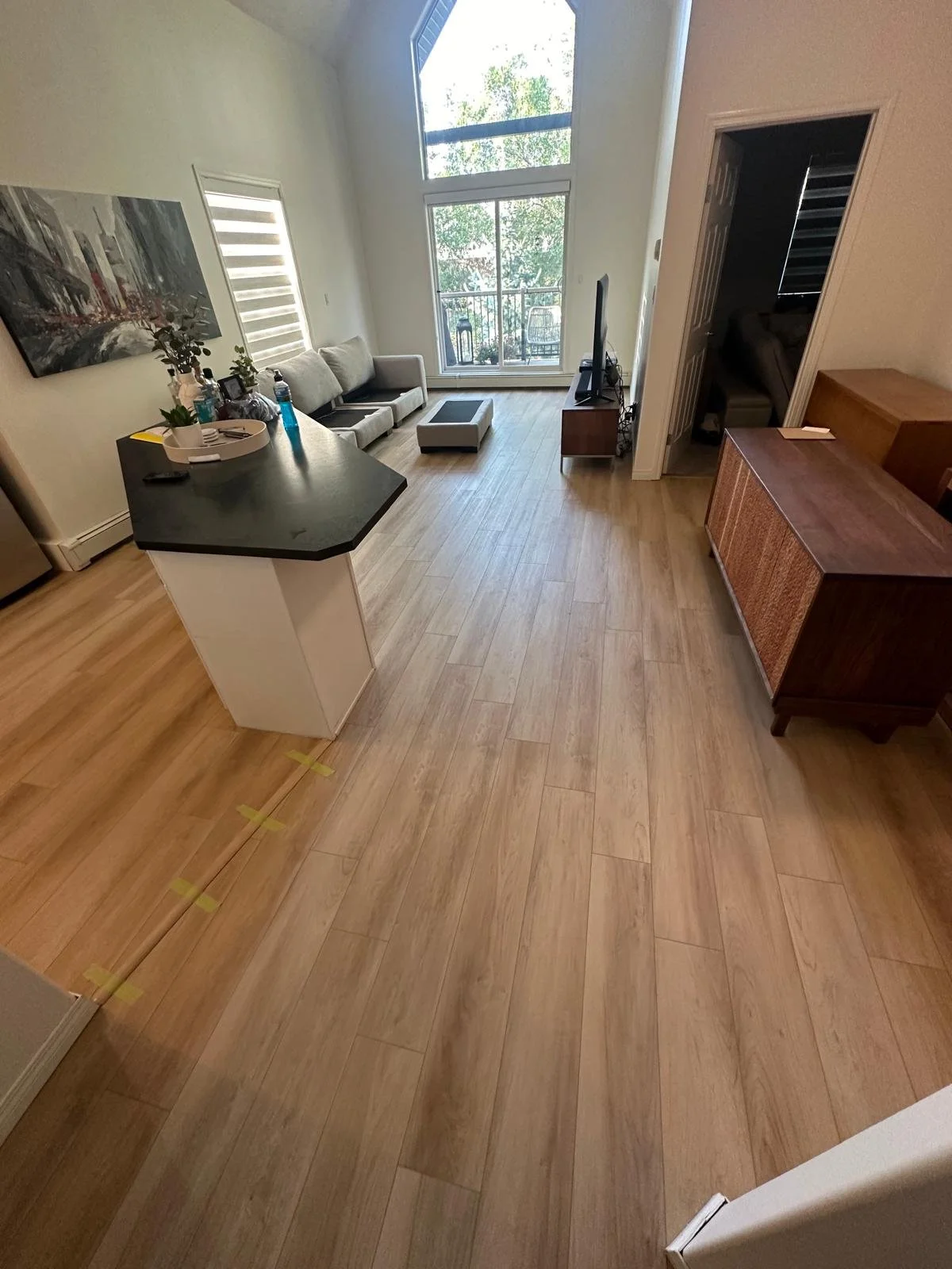 Spacious modern interior with large windows, wooden floors, and people sitting against the wall.